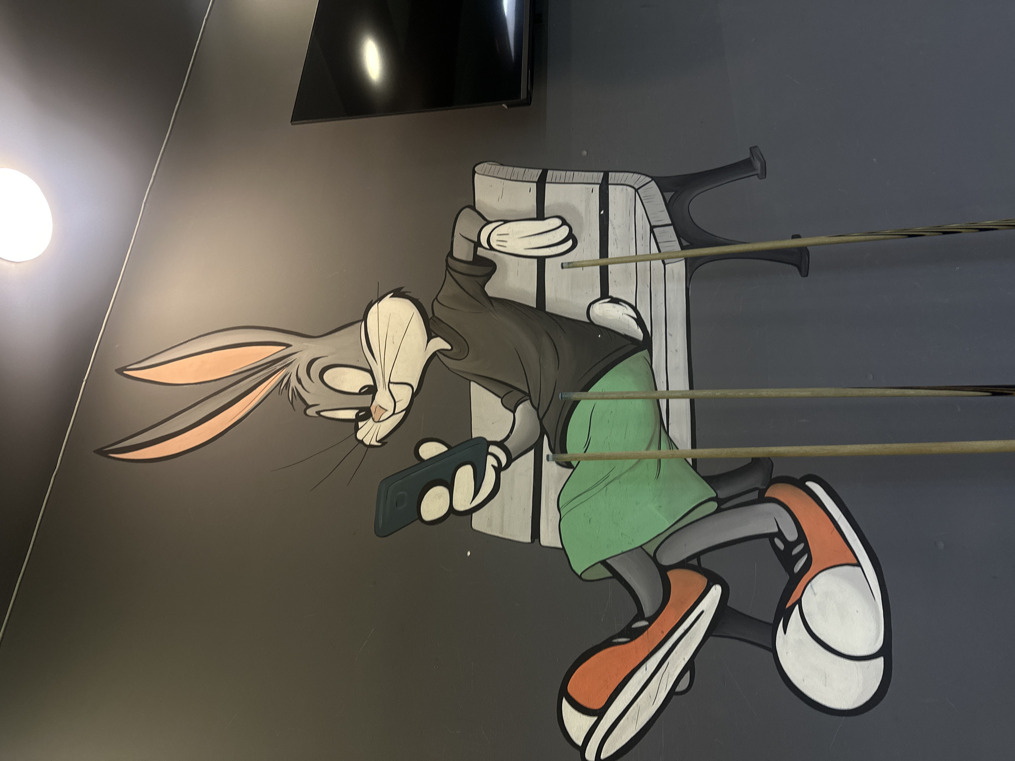 A mural on a gray wall in Yerevan, Armenia, depicts a cartoon figure resembling Bugs Bunny. The figure is portrayed as being struck by billiard cues, with three cues visually piercing through the character's body. The figure is holding a black smartphone and wears orange and white sneakers, a green skirt, and a gray top. A flat-screen television is mounted on the wall above the mural. A ceiling light fixture is visible in the upper-left corner.