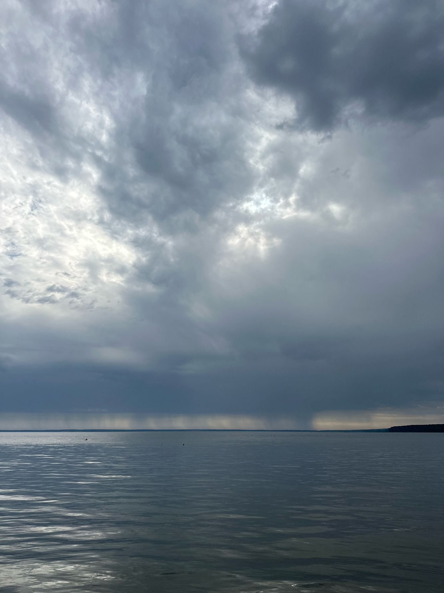 The image depicts a wide expanse of calm water under a dramatic, overcast sky. The water's surface is rippled, reflecting the muted light filtering through the thick clouds. In the distance, a faint line of dark land marks the horizon. A thin veil of rain is visible falling in streaks over a section of the water. The sky is filled with layers of gray clouds, with some brighter patches where sunlight is attempting to break through. There are no discernible people or specific structures in the scene. The overall atmosphere is one of impending weather or a recent shower.