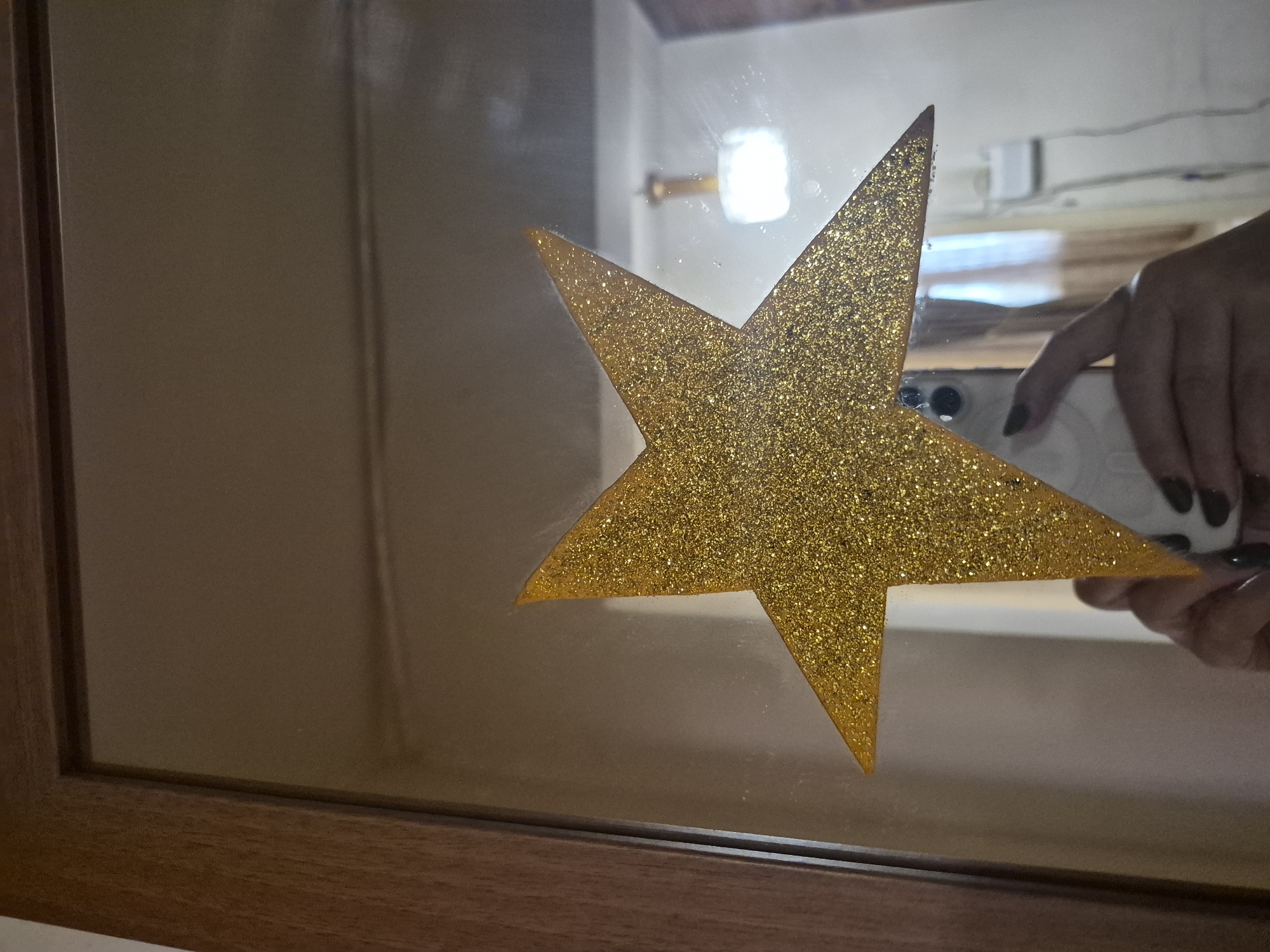 A close-up shot shows a large, golden, glitter-covered star adhered to a mirror. The star occupies the center of the frame and reflects the surrounding environment. In the reflection, a person's hand is visible holding a smartphone, suggesting they are taking the photograph. A lamp with a square shade is also visible in the reflection, as well as a white electrical outlet with cords. The mirror appears to be framed by dark wood, possibly part of a door or furniture. The lighting is soft and diffused. There is no discernible context that places this image specifically in Yerevan, Armenia.