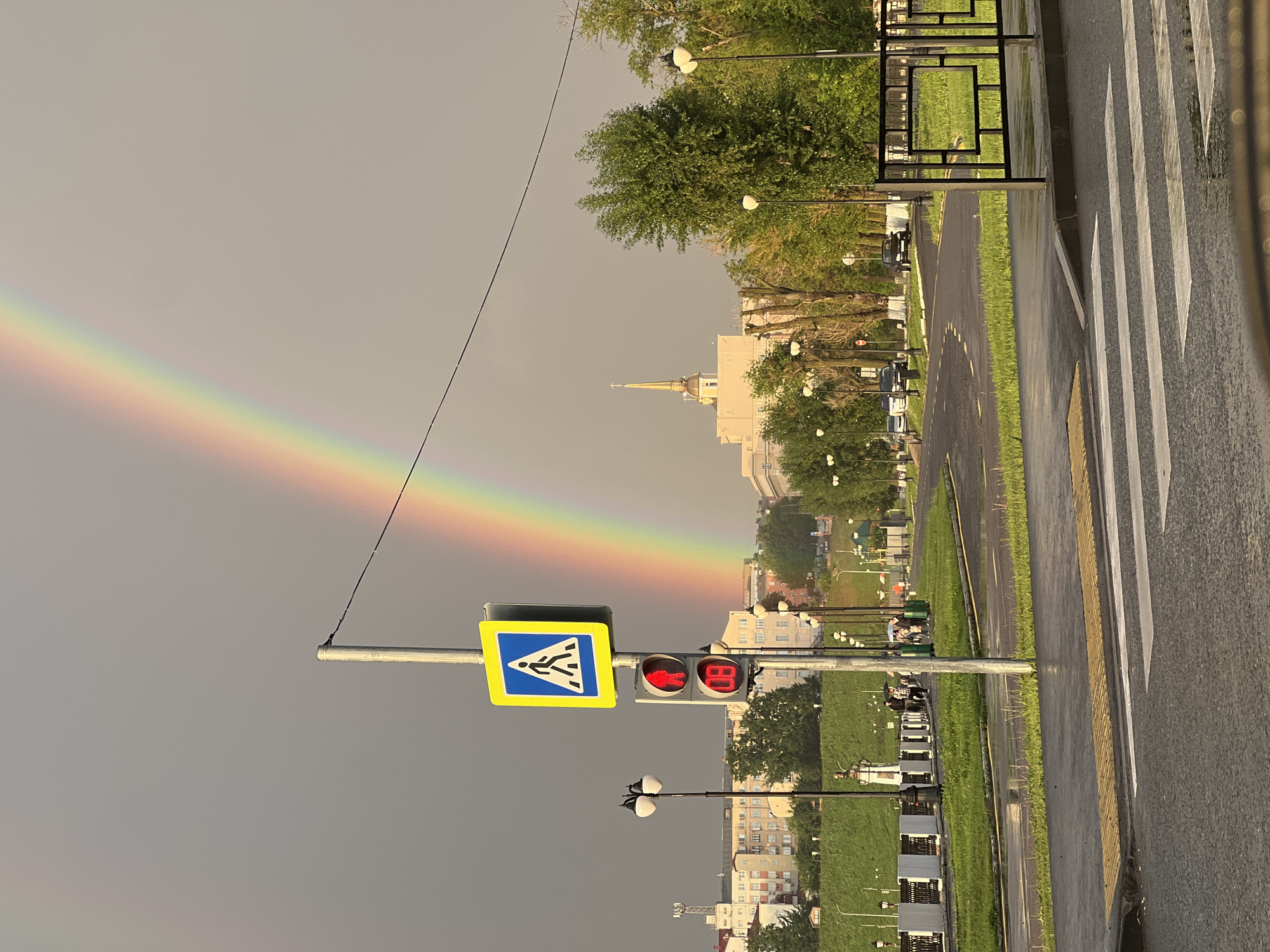 The image captures a scene in Izhevsk, Russia, with a prominent rainbow arcing across a gray, overcast sky. The perspective is tilted, making the ground appear vertical. In the foreground, a pedestrian crossing with white stripes is visible on a wet asphalt road. Adjacent to the crossing is a traffic light displaying a red signal for pedestrians and a digital display showing "80". A blue pedestrian crossing sign with a yellow border is mounted on the same pole as the traffic light.
Beyond the road, a strip of green grass separates it from a paved path lined with trees. Streetlights with white globes are spaced along this path. In the background, there are multi-story buildings, some with light-colored facades. A building with a golden spire, possibly a church or administrative building, stands out in the mid-ground. The presence of the wet road and the subdued lighting suggest recent rain and a possibly humid atmosphere. The rainbow indicates a meteorological phenomenon occurring during or after rainfall with sunlight. No people are clearly identifiable in the image, though a few figures can be discerned from a distance on benches along the path. The overall mood is serene, with the natural beauty of the rainbow juxtaposed against the urban environment. The visible text on the traffic signal is "80".