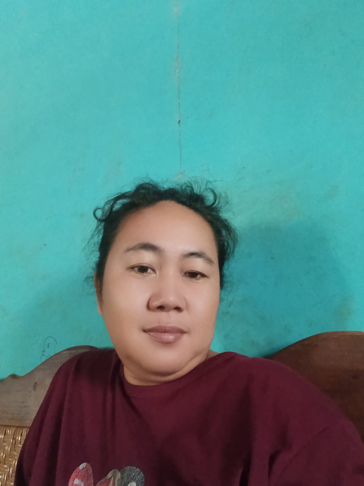 A head and shoulder shot of a woman is captured from the chest up. She is facing forward, looking directly at the camera. Her dark hair is pulled back loosely, with some strands falling around her face. She is wearing a maroon-colored t-shirt with a graphic design on the front. Behind her, a turquoise wall serves as the backdrop, with a visible seam running vertically down the center. To the right, a portion of a wooden chair with woven detailing is visible. The image appears to have been taken indoors, possibly in a residential setting in Karangmojo, Indonesia.