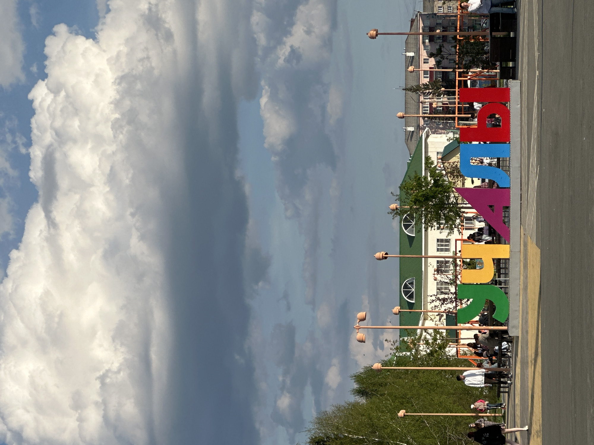 The image shows a sunny urban scene. In the foreground, there's a large, colorful sign spelling out what appears to be a place name in Cyrillic letters, likely representing the city or region. People are casually walking and sitting on or around the sign, suggesting it is a popular gathering spot or public art installation. Behind the sign, there is a street with lampposts and buildings, including a green-roofed structure with arched windows. Further back, there are more buildings, some appearing residential. The sky occupies a significant portion of the image, filled with dramatic, textured clouds, ranging from bright white to dark gray. The image is taken during the daytime with good lighting.