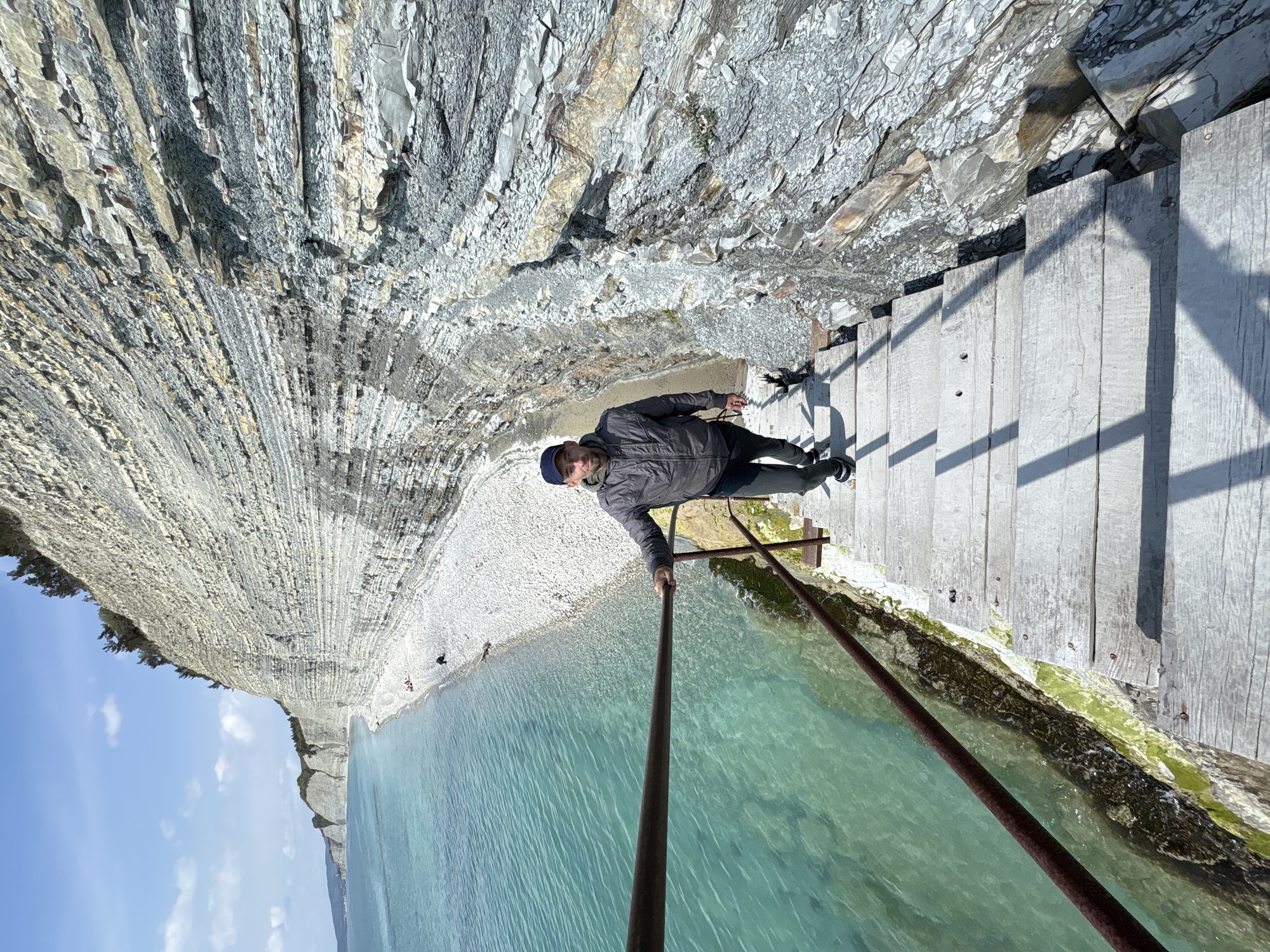 This daytime image, taken in Gelendzhik, Russia, captures a man on a wooden walkway built along a dramatic coastal cliff. The scene is bright and sunny, with a clear blue sky and a few scattered clouds.
The main subject is a man, dressed in a dark jacket, dark trousers, and a dark cap, standing on the wooden platform. He is holding onto a rusty metal handrail that separates the walkway from the water. He appears to be posing for the camera, looking towards it with a neutral to slightly smiling expression.
To the man's left, a towering, layered rock cliff dominates the landscape. Its distinctive geological formation shows numerous horizontal and slightly curved strata, giving it a textured, almost corrugated appearance. The cliff face exhibits various shades of grey, white, and light brown rock.
To the man's right, beyond the metal railing, lies a body of remarkably clear, turquoise-colored water, likely the Black Sea. Patches of green algae are visible on the submerged parts of the structure beneath the walkway. In the background, further along the coast, a pebble beach stretches at the base of the cliffs, with a few distant figures visible. More cliffs extend into the horizon under the bright sky.
The wooden walkway itself is made of light-colored planks, and the metal railing casts distinct shadows across it, indicating the bright sun. The impressive scale of the cliffs and the pristine coastal waters are notable features of this location. No visible text is present in the image.