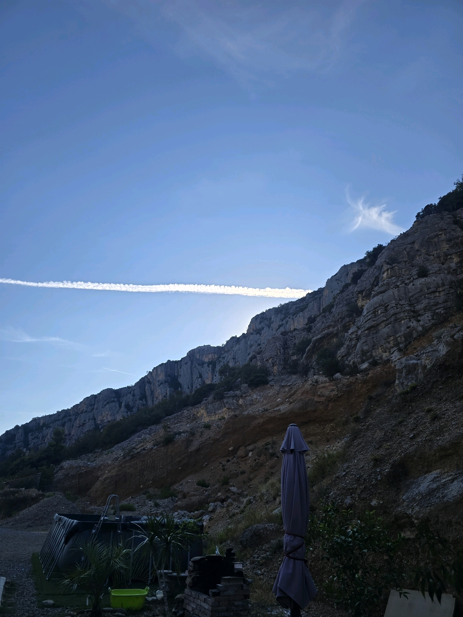 This media file captures a serene outdoor scene, likely in Podgorica, Montenegro, during the late afternoon or early evening. The dominant feature is a rugged, rocky mountainside that occupies the right two-thirds of the frame, sloping upwards towards the sky. The mountain face is textured with layers of rock and sparse vegetation.
In the sky, a long, thin contrail stretches horizontally across the frame, indicative of an aircraft having recently passed overhead. Scattered wispy clouds are also visible, particularly a more elaborate, feathery cloud formation in the upper right quadrant. The sky itself transitions from a clear, bright blue near the horizon to a deeper shade towards the top.
In the foreground, on the left side, a small, above-ground swimming pool is partially visible. Next to it, a closed, dark gray umbrella stands upright, tied with rope. Several potted plants and a bright green bucket are also scattered in the foreground, suggesting a residential or recreational area. The overall mood of the image is peaceful and tranquil, enhanced by the soft, diffused light. The scene appears to be set in a somewhat undeveloped or natural area, given the presence of the rocky mountain and the rustic elements in the foreground. No people are visible in the frame, and no text is present.