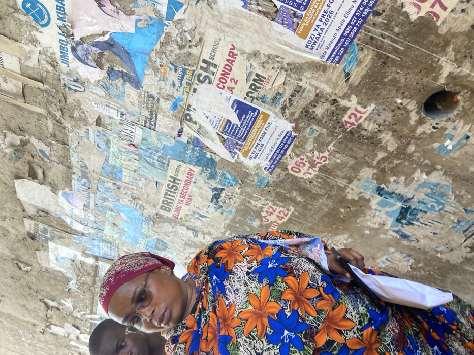 An eye-level shot captures a crowded, aged wall plastered with faded school advertisements. The posters, partially torn, promote British School, secondary education, and form enrollment. Text such as "Jimbo Kiba" and phone numbers are visible. The lower part of the image features a woman with glasses and a patterned headscarf and dress. Her head is slightly bowed, and she holds a white envelope or document. A portion of another person's head is seen. The overall impression is a busy urban street scene with an emphasis on education opportunities.