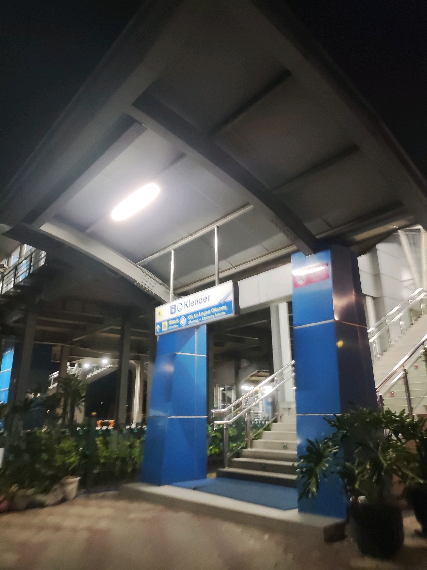 The image captures the exterior of the Klender station, an elevated train station located in East Jakarta, Indonesia, at night. The station features a modern architectural design with a prominent blue pillar and a set of stairs leading up to the platform. A sign indicates "Klender" as the station name, with additional text in Indonesian mentioning train lines and destinations. The station is illuminated by overhead lights, casting a glow on the surroundings. Potted plants are visible at the base of the stairs, adding a touch of greenery to the urban landscape. The overall scene depicts a well-maintained public transportation hub during nighttime hours.
