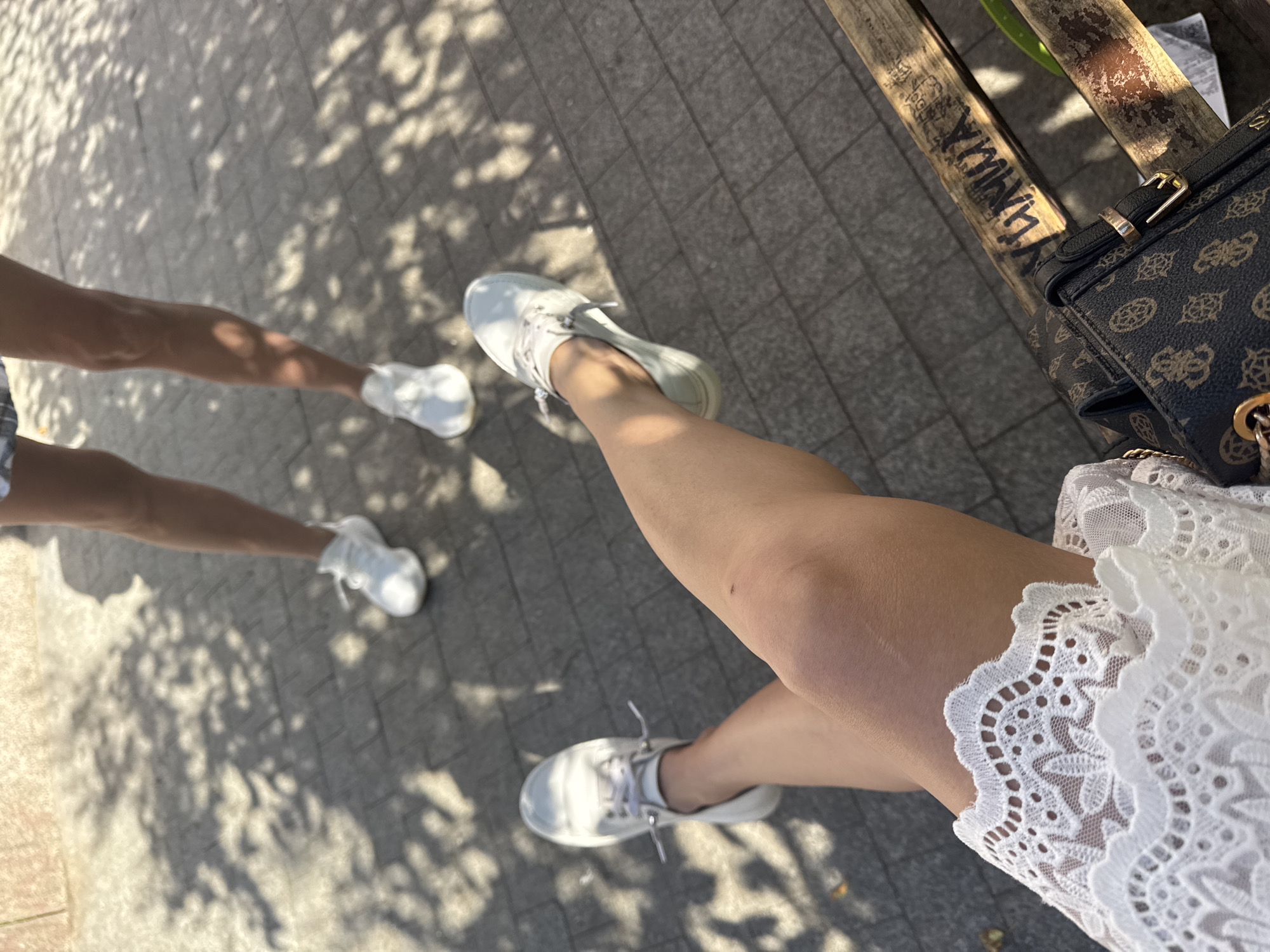 The image shows two individuals, likely females, in the city of Saint Petersburg, Russia. The foreground features a person's legs and feet, adorned with white sneakers and a white lace top. They are seated on a wooden bench, with a patterned handbag beside them. The background displays another person's legs and feet, also wearing white sneakers, standing on a paved walkway. Sunlight casts dappled shadows from overhead foliage onto the ground, suggesting an outdoor setting in a park or public square. The overall scene depicts a casual, leisurely moment, possibly during daytime.