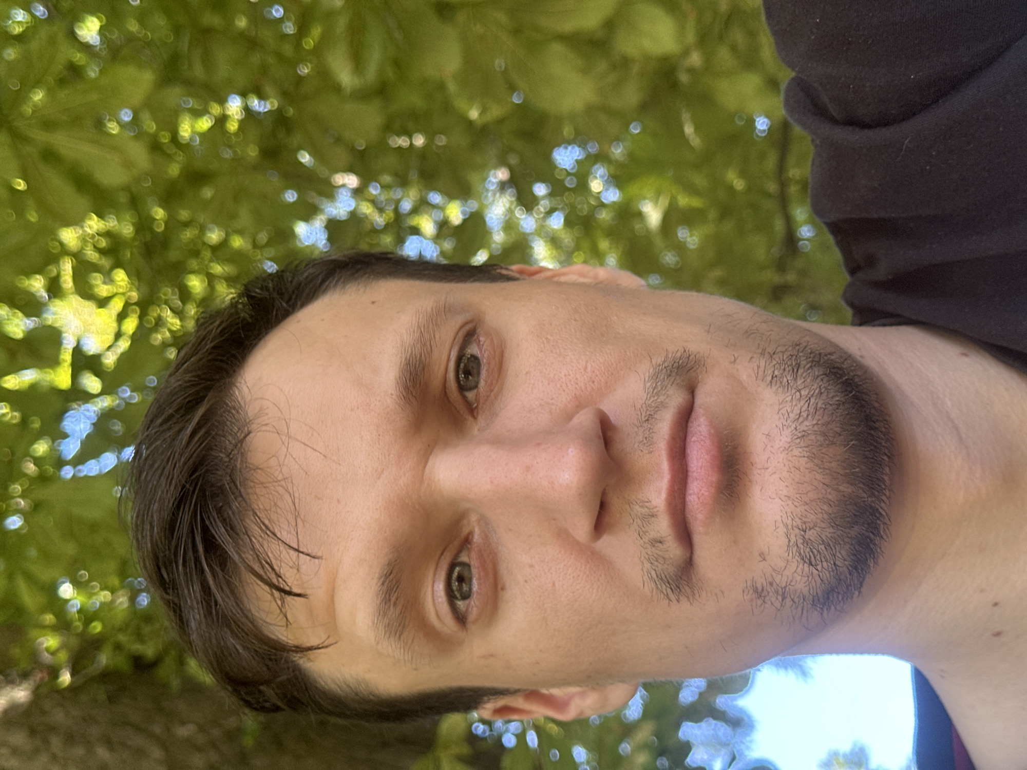 This is a close-up, selfie-style photograph of a man taken outdoors with a blurred green leafy background. The man, who appears to be an adult male, is facing the camera. His head and upper torso are visible. He has dark hair, light skin, and stubble on his chin. His expression is neutral. The lighting suggests it is daytime, possibly with sunlight filtering through the leaves, creating bokeh effects. There is no indication of the location being in Makiv, Ukraine, or any other specific city. The image focuses solely on the man's face and the immediate foreground.