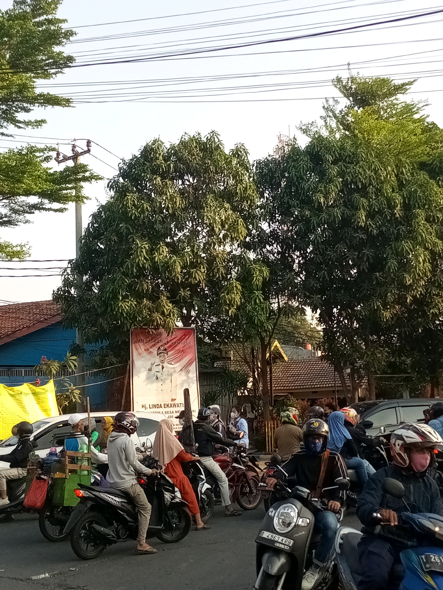 The image depicts a street scene in Tambun, Indonesia, during what appears to be late afternoon, given the soft lighting. The foreground is dominated by a group of motorcycles and their riders, some stationary in traffic, others moving. Several individuals are wearing helmets and face masks, common precautions in the region. One motorcycle has a green cart attached to the back, loaded with goods. Behind the motorcycles, a large banner is prominently displayed, featuring a portrait of a person in official attire, likely a political figure, with Indonesian text visible, suggesting an election or campaign event. Beyond the banner, trees with lush green foliage create a canopy, with some buildings and power lines also visible. The overall impression is one of daily life in a busy Indonesian town.