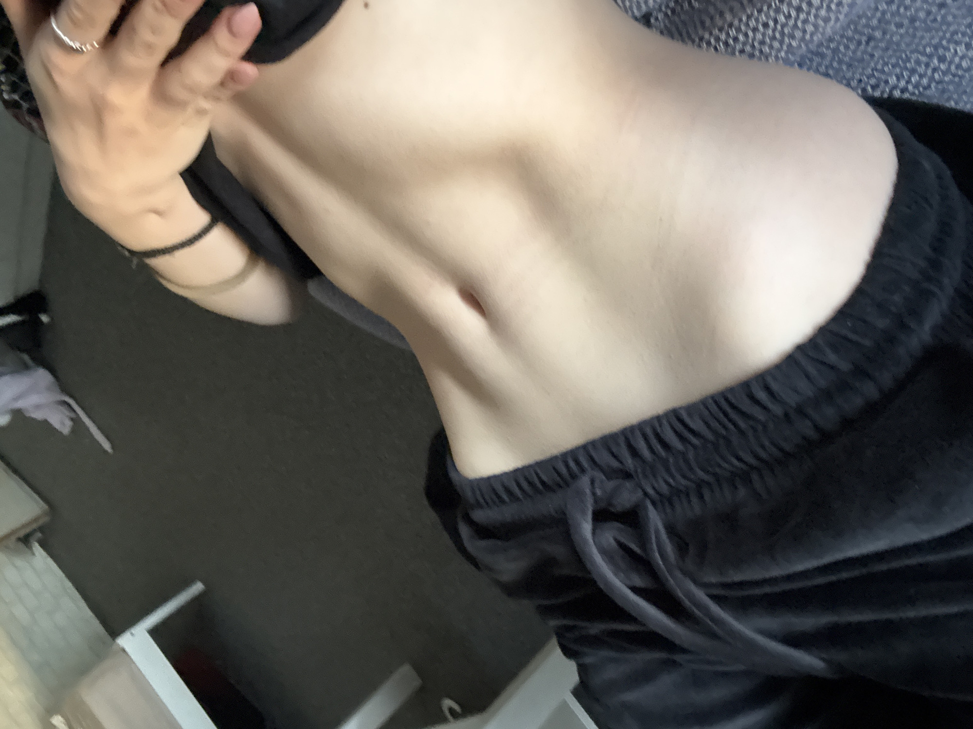 The image depicts a person's torso from the upper chest down to the upper thighs, wearing a black bra and dark velvet sweatpants. The person is standing indoors, and the background includes part of a room with a carpeted floor and a white shelf unit. A hand is positioned near the chest area. There is no information available in this image to indicate that the location is Sofia, Bulgaria.
