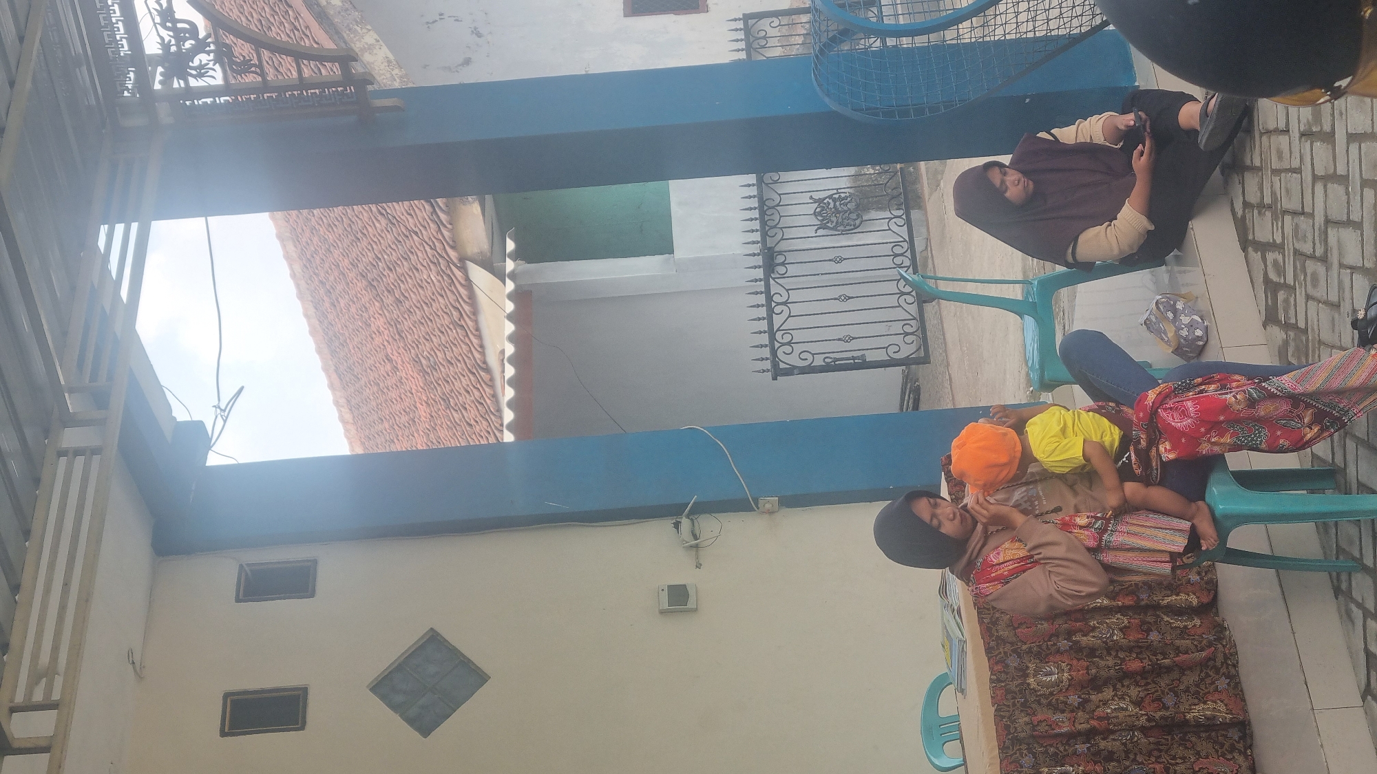 In Diwek, Indonesia, a mother, wearing a dark hijab and a light brown hooded sweatshirt, sits on a turquoise plastic chair holding a young child. The child, dressed in a yellow t-shirt and orange beanie, is held in her arms, wrapped in a colorful textile. To the right, another woman in a dark hijab and a beige sweater sits on the ground, looking down at her smartphone. In the background, a wrought iron gate separates the paved outdoor area from a building with a red-tiled roof. The scene appears to be a casual gathering or waiting area, possibly at a public space or residence, under a covered patio with blue support beams.