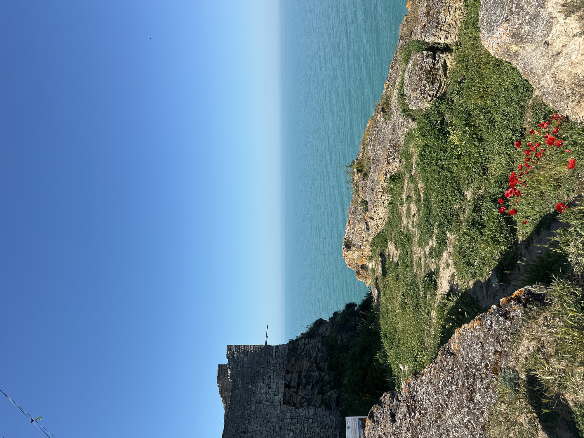 The image depicts a view from a cliffside overlooking the sea, in Balgarevo, Bulgaria. The sky is clear blue. The sea is a gradient from darker to lighter blue towards the horizon. On the right side of the frame, a rocky cliff is covered with green grass and patches of red poppies. The cliff edge appears uneven and natural.
On the left side, there is a partially ruined stone structure or wall. The construction is old, with stones fit together to form a wall. The image is taken from an elevated perspective, looking down onto the sea and across to the horizon.