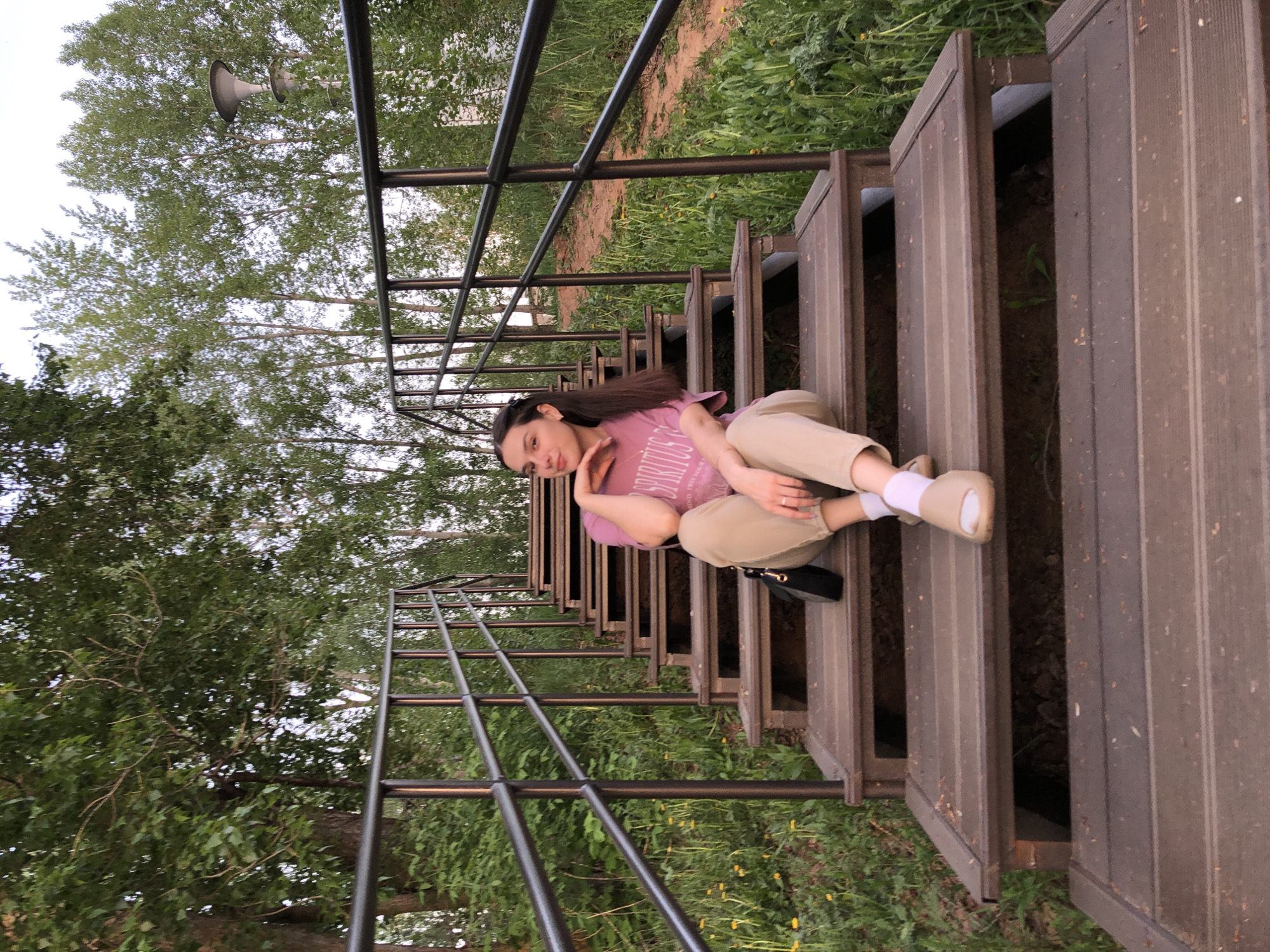 A young woman with long dark hair is sitting cross-legged on a wooden staircase in what appears to be a park or outdoor setting. She is wearing a light purple t-shirt, beige pants, white socks, and light-colored sandals. A black purse is resting on the step beside her. The staircase has metal railings on either side. Lush green foliage and trees surround the staircase. Based on visual similarities, this location could be in the city of Chaikovsky, Russia.