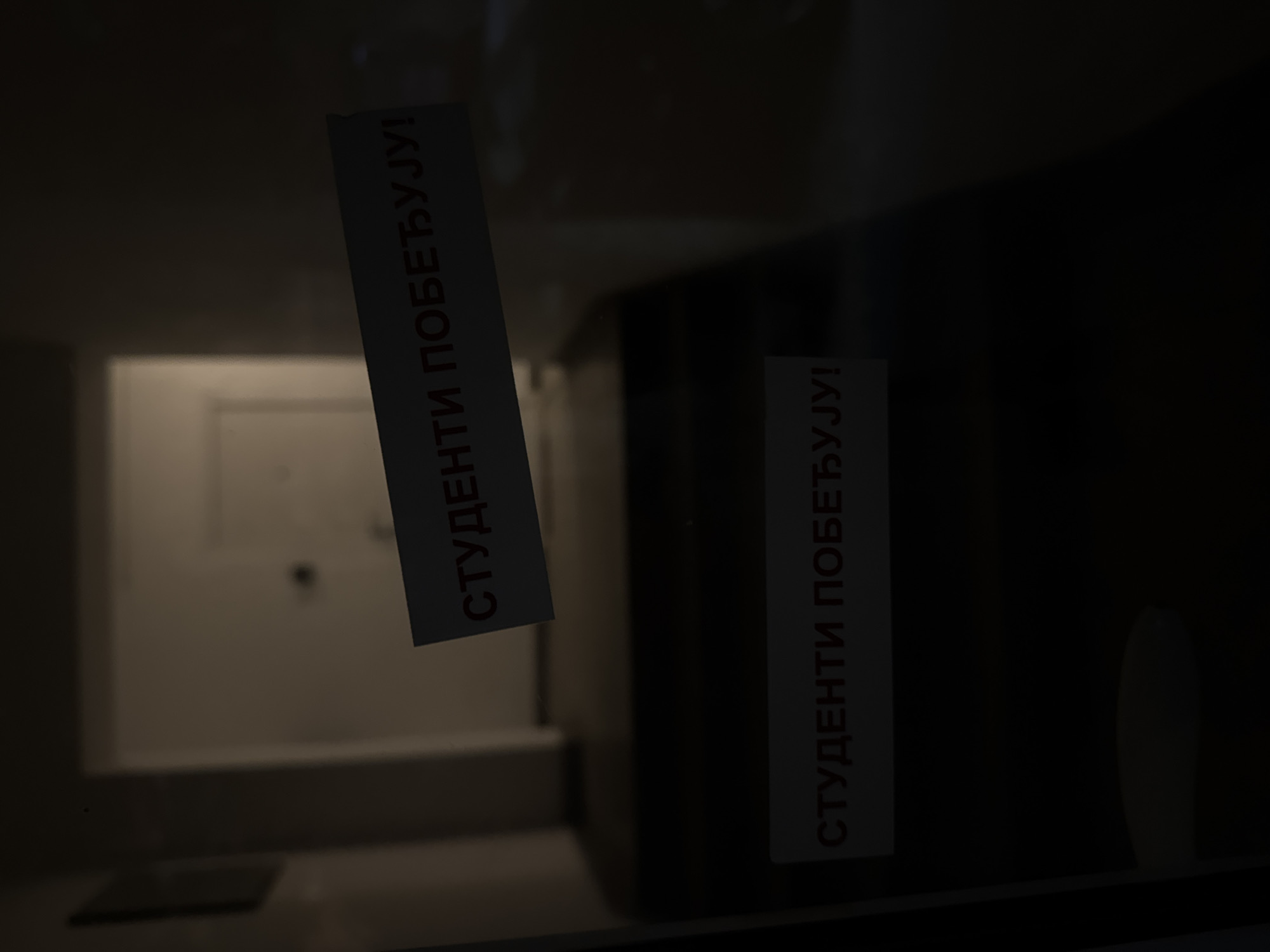 The image shows a dark hallway with two signs on what appears to be a glass door or partition. Both signs are white with red Cyrillic lettering. The top sign reads "СТУДЕНТИ ПОБЕЂУЈУ!". The bottom sign, positioned lower down, reads "СТУДЕНТИ ПОБЕЂУЈ!". The hallway has a set of dark stairs leading up into the darkness. The environment is dimly lit, suggesting it might be nighttime or an area with poor lighting. No people are visible in the frame.