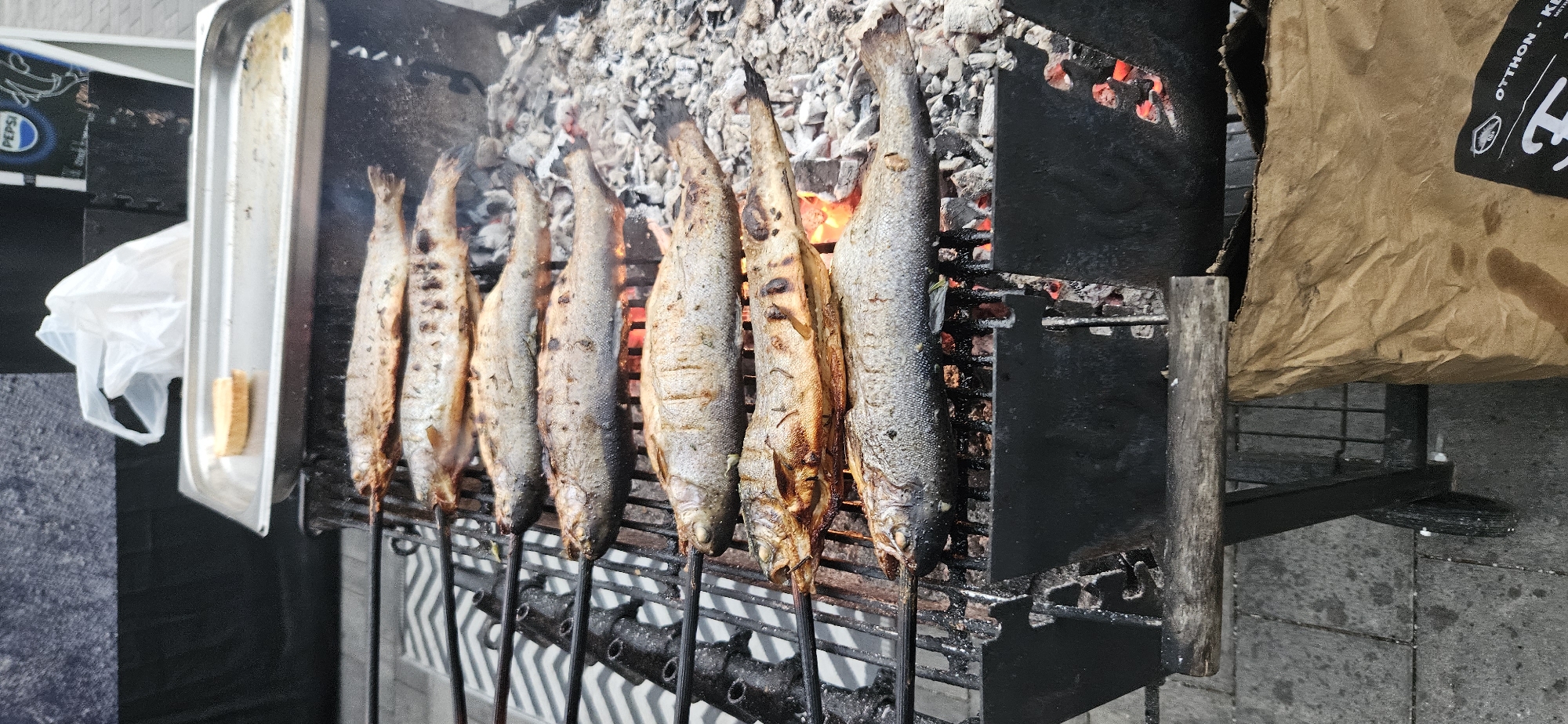 The image shows whole fish grilling on a charcoal barbecue in Hévíz, Hungary. Seven fish are visible, skewered and arranged on a metal grate over glowing coals. The fish appear to be seasoned and are in various stages of cooking, with some exhibiting char marks. Above the grill, a stainless steel tray holds a piece of bread. The background includes a black cloth barrier, a white plastic bag, and a partial view of what appears to be a Pepsi advertisement. The barbecue itself is situated outdoors on a paved surface, with a brown paper bag visible in the lower right corner. The overall scene depicts an outdoor food preparation setting.