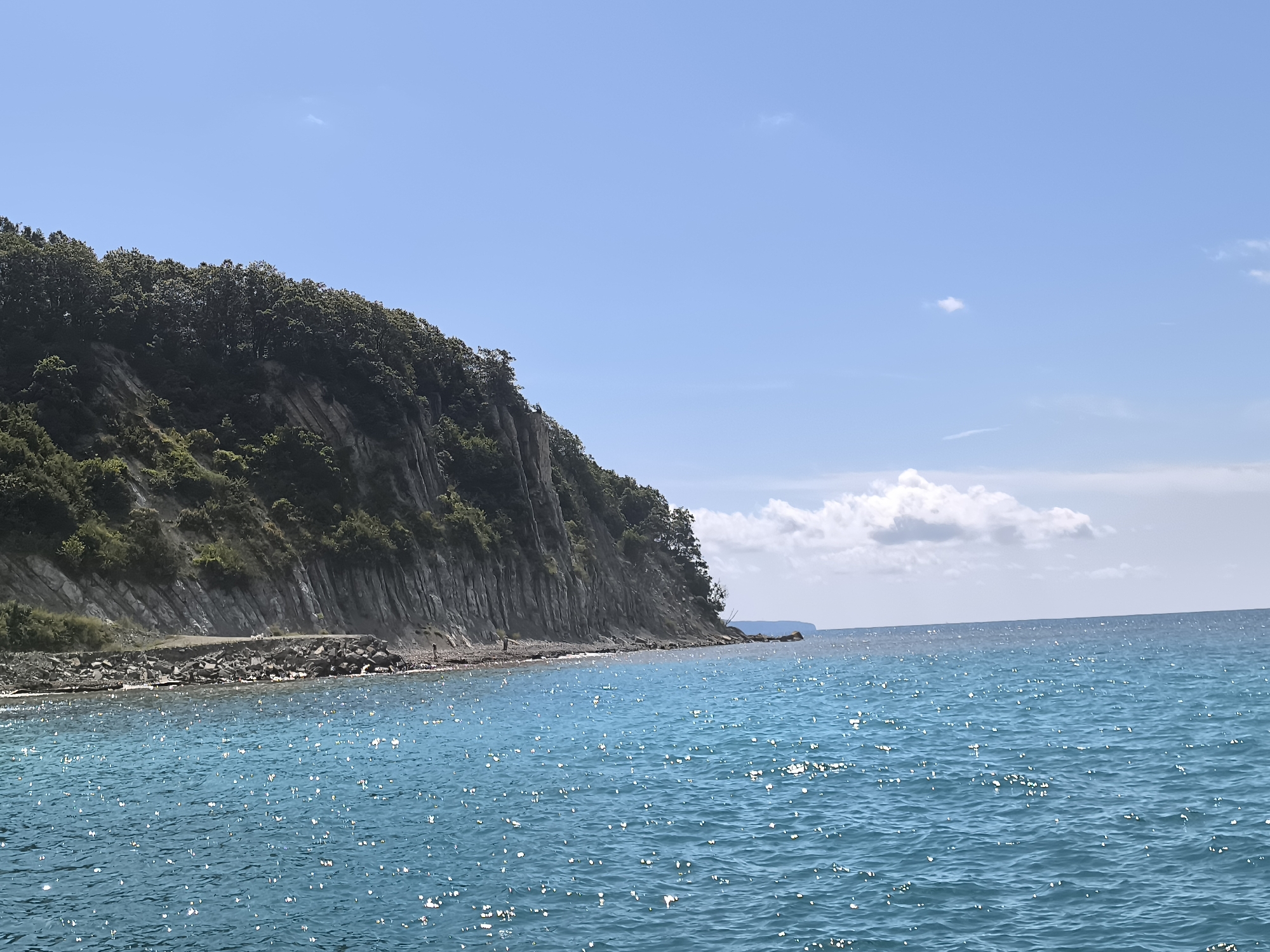 The image captures a scenic coastal view, likely near the city of Lermontovo, Russia. A steep, tree-covered cliff dominates the left side of the frame, meeting the turquoise waters of the sea. The cliff face appears rocky with visible strata. Along the base of the cliff, a narrow strip of rocky shore is visible, with a few small figures appearing to be standing or walking on it. The sea is a vibrant blue, with sunlight glinting off its surface, creating a shimmering effect. A few white clouds dot the clear blue sky. The overall impression is one of a tranquil natural landscape.
