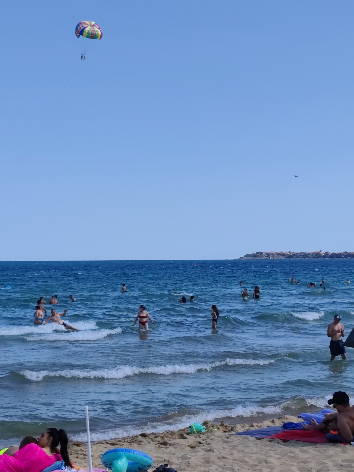 The image depicts a sunny day at a crowded beach in Nesebar, Bulgaria, with the ancient town visible on the peninsula in the distance. The turquoise sea is active with moderate waves, and numerous people are enjoying the water. In the sky, a colorful parachute with two individuals is being towed by a boat, a common sight for water sports enthusiasts.
In the water, a diverse group of people are swimming, wading, and playing. Some are closer to the shore, while others venture further out. A man in a red bikini is standing, and a man with dark hair is partially submerged. Several other individuals are visible in various states of activity within the sea.
On the sandy beach in the foreground, several people are relaxing. A woman wearing a bright pink garment is lying down, and a child is next to her. A blue inflatable ring is placed on the sand, and a man wearing a black baseball cap is seated on a mat, looking down at something in his hands.
The sky is clear and blue, with a bright sun illuminating the scene. A small bird is captured in flight in the upper right portion of the image. The overall atmosphere is one of leisure and summer enjoyment.