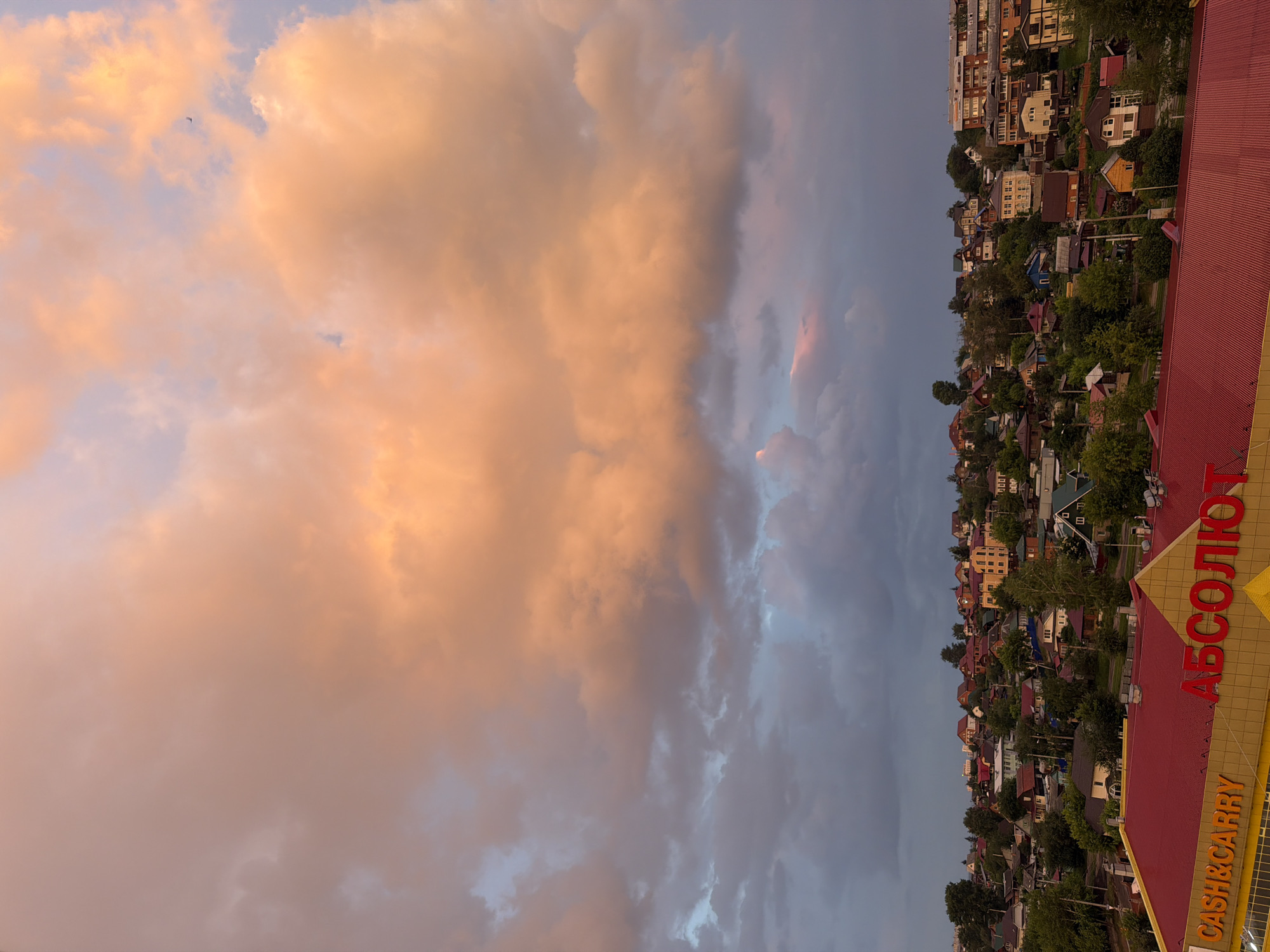 The image depicts a panoramic view of a city nestled on a hillside, with a prominent commercial building in the foreground. The sky above is dramatic, filled with large, fluffy clouds illuminated by the warm hues of a sunset, ranging from soft peach to vibrant orange.
In the foreground, the red roof of a commercial building dominates the lower portion of the frame. Yellow signage with orange lettering is visible, with "CASH&CARRY" on the left and what appears to be "АБСОЛЮТ" (Absolut) on the right, indicating a retail establishment.
Beyond the commercial building, the hillside is densely populated with a variety of houses, ranging from single-family homes to multi-story apartment buildings. The architecture suggests a mix of traditional and more modern designs, with many houses featuring colorful roofs and gardens. Lush green trees are interspersed throughout the residential area, adding to the natural beauty of the scene.
The city appears to be located in Irkutsk, Russia, as suggested by the Cyrillic lettering on the signage. The time of day is clearly sunset, with the warm light casting a serene and picturesque glow over the urban landscape. There are no visible people or animals in the image. The overall impression is one of a bustling city at dusk, bathed in the warm, colorful light of the setting sun.