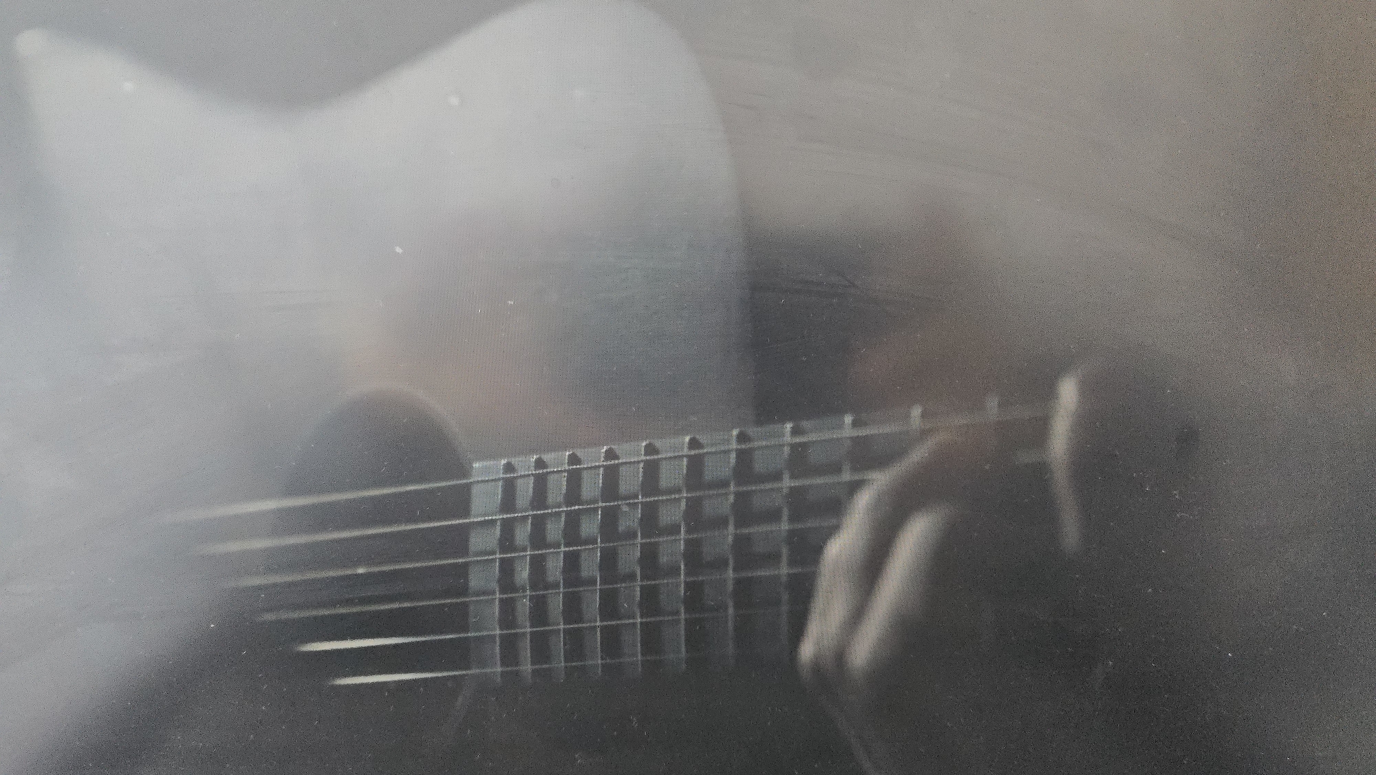 A hazy, monochrome image depicts a person playing a guitar. The hand is visible pressing down on the strings on the fretboard. The guitar's body and neck are also partially visible, although obscured by the haze. No specific city or location can be determined from the image.