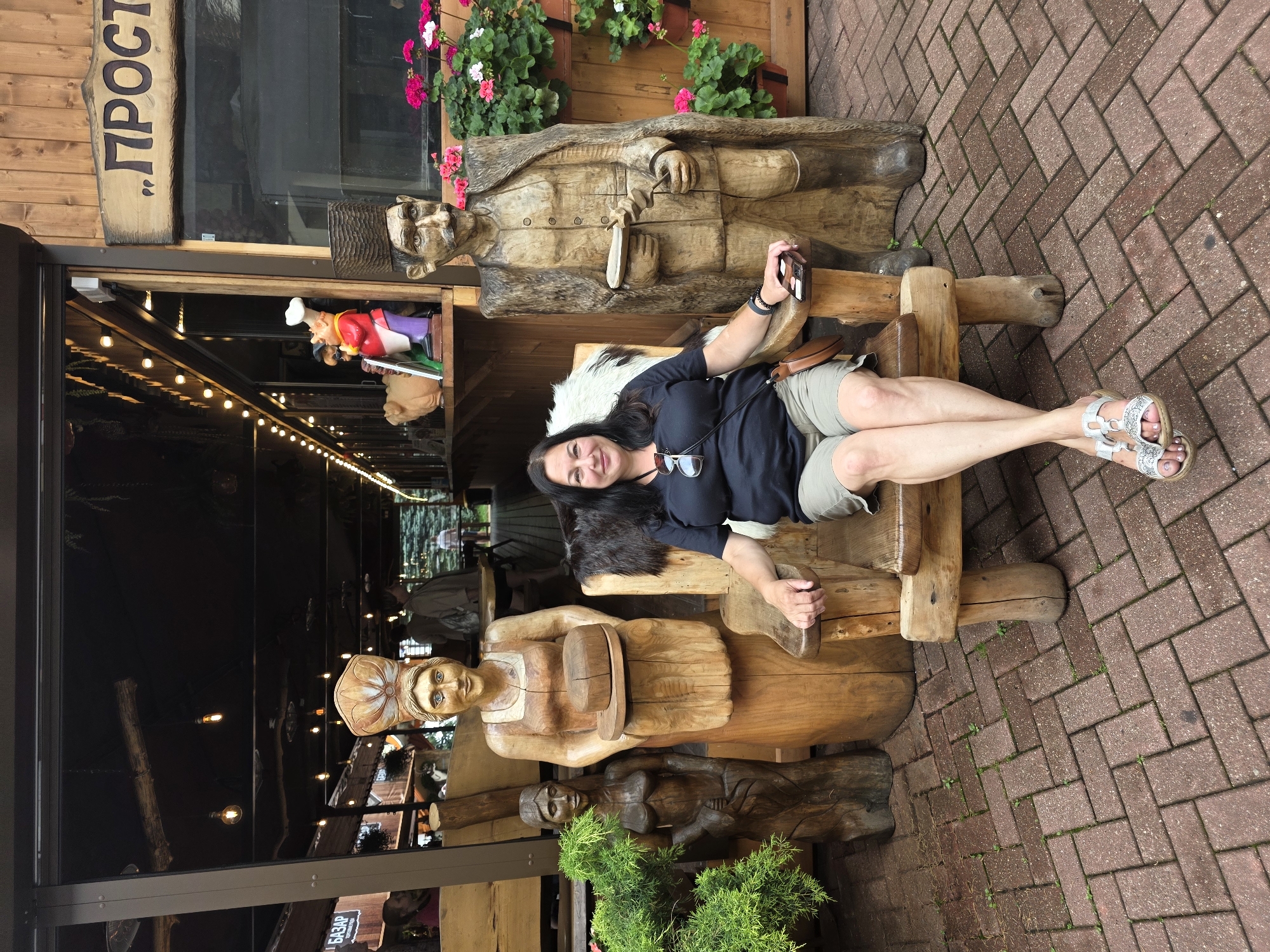 A middle-aged woman with dark hair sits on a large, carved wooden chair in front of a wooden building. She is wearing a dark t-shirt and khaki shorts, and silver sandals. Her legs are crossed, and she is holding a mobile phone in her right hand. To her left is a carved wooden statue of a woman holding a loaf of bread. To her right is a carved wooden statue of a man wearing a fur cloak and a tall hat. The building behind them has a sign with Cyrillic lettering that reads "ПРОСТО". There are potted plants with pink and white flowers to the right of the man statue. The ground is paved with red brick pavers. The scene appears to be outdoors, possibly a cafe or restaurant, in the city of Estosadok, Russia.