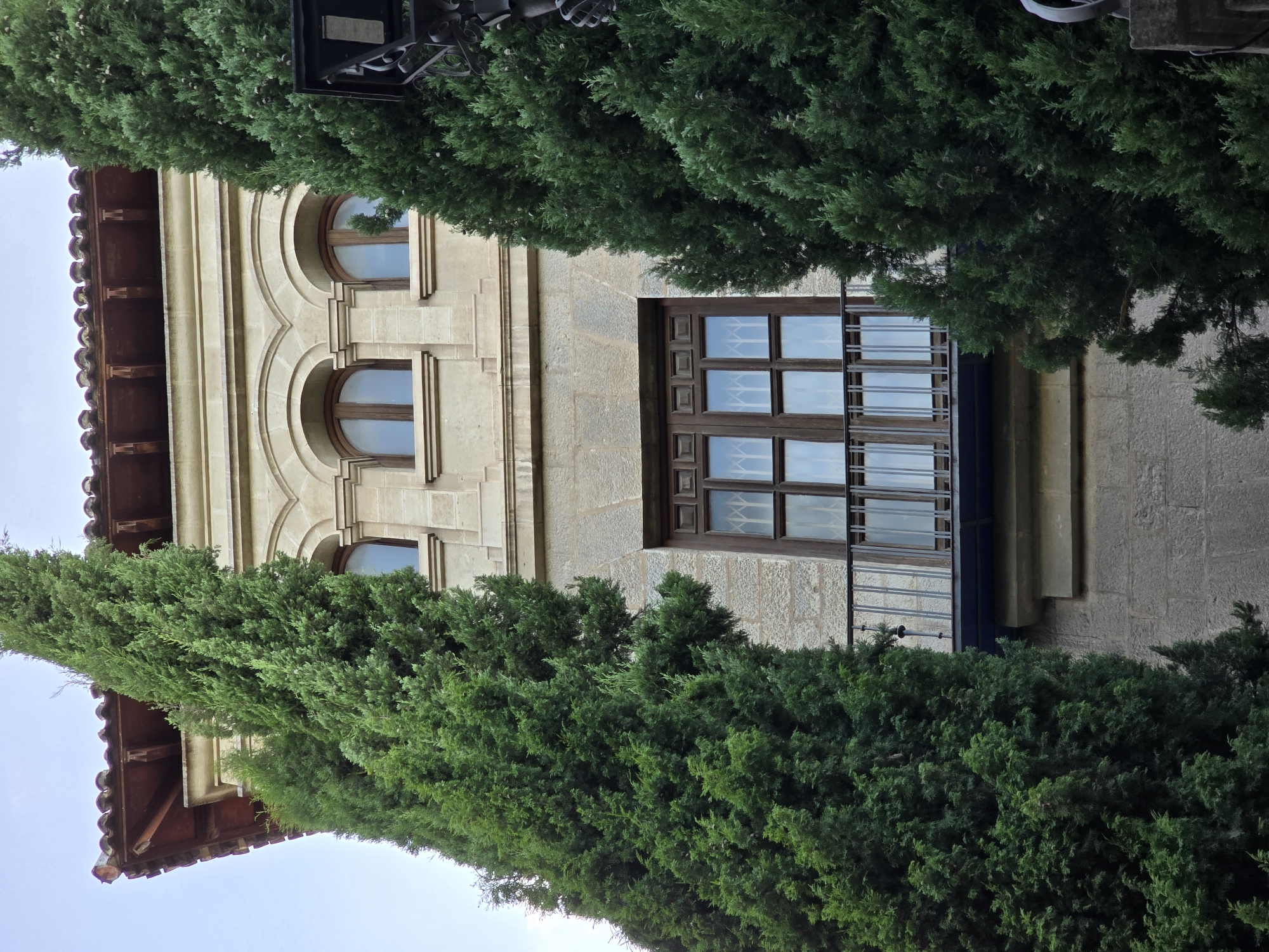 This is a low-angle, medium shot of the exterior of a stone building with a tiled roof, partially obscured by two tall, dense green cypress trees. The building features several arched windows on the upper floor and a large, multi-paned window with a dark wooden frame and a wrought-iron balcony on the lower section. The sky is visible in the upper portion of the image, appearing slightly overcast. The building's architecture suggests a historical or traditional style. The scene is devoid of any people or specific actions. The location is a city in Jaén, Spain.