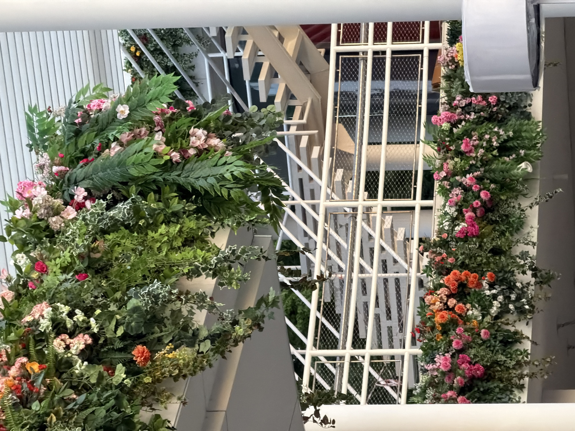 The image depicts a floral decoration in what appears to be an indoor setting, possibly a building interior. A lush arrangement of foliage and flowers, featuring pink, orange, and white blooms, is draped around white architectural elements, including a ladder-like structure and vertical columns. The scene suggests an event or decorative installation. There is no clear indication of the location being Moscow, Russia.