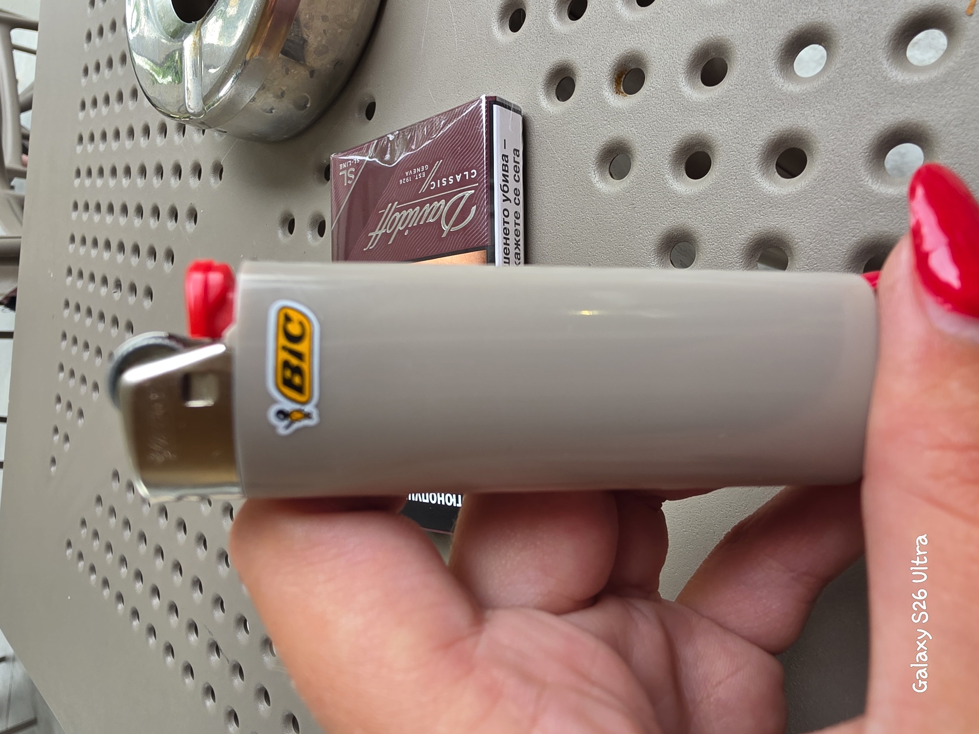 A person's hand with bright red fingernail polish is holding a light brown Bic lighter on a tan textured table. A pack of Davidoff Classic cigarettes and a metal ashtray are visible on the table to the right of the lighter. The table surface has a pattern of circular indentations. Text on the cigarette pack is partially obscured, but includes "Davidoff CLASSIC GENEVA" and Cyrillic text. A watermark in the bottom left corner reads "Galaxy S26 Ultra." The scene is likely in an outdoor seating area in Plovdiv, Bulgaria, given the context provided.