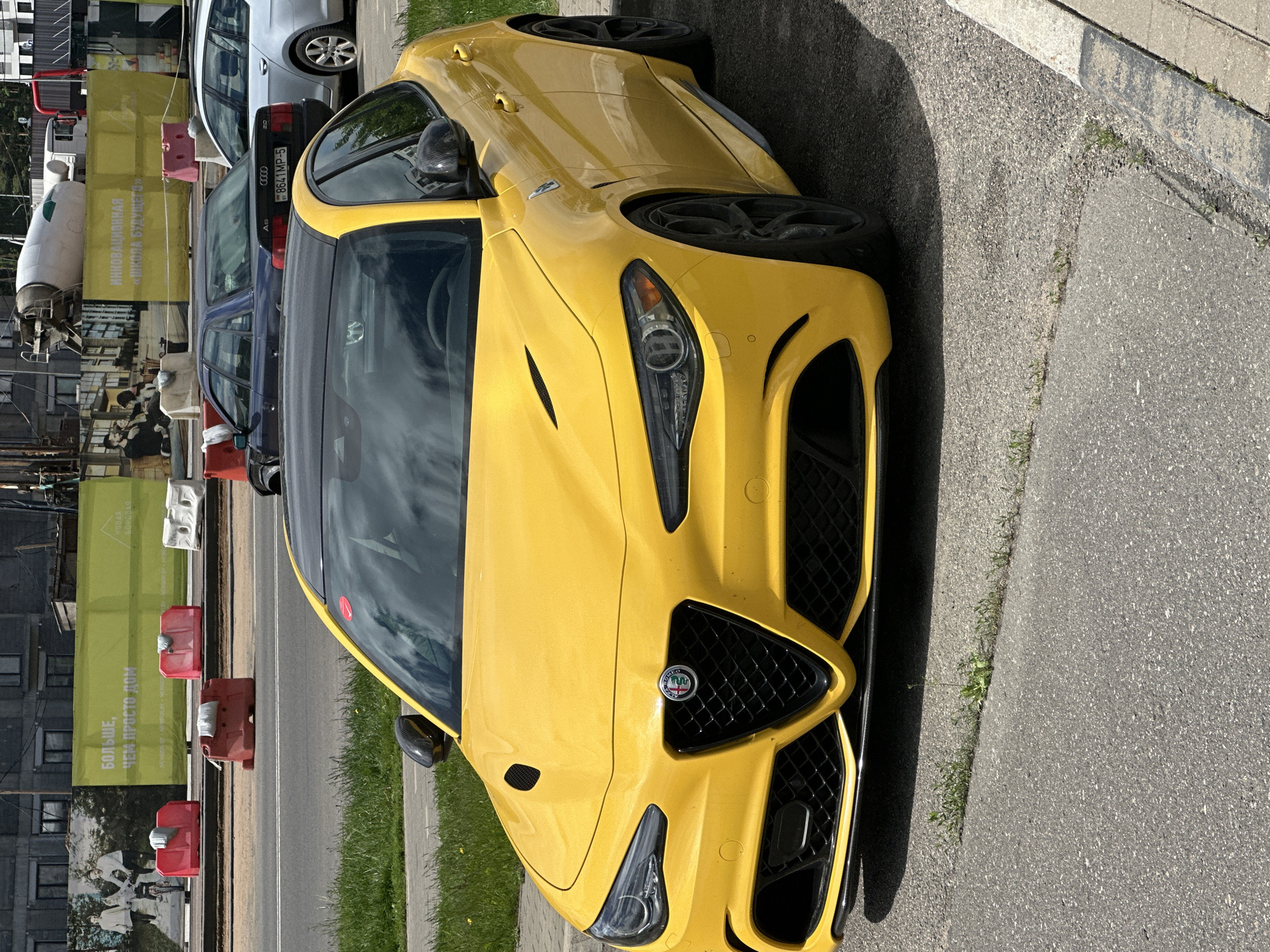 The image shows a yellow Alfa Romeo Giulia parked on the side of a road in Бараўлянскі сельскі Савет, Belarus. Other cars are also parked nearby, including a silver Audi. The background includes buildings, signs, and construction materials.
