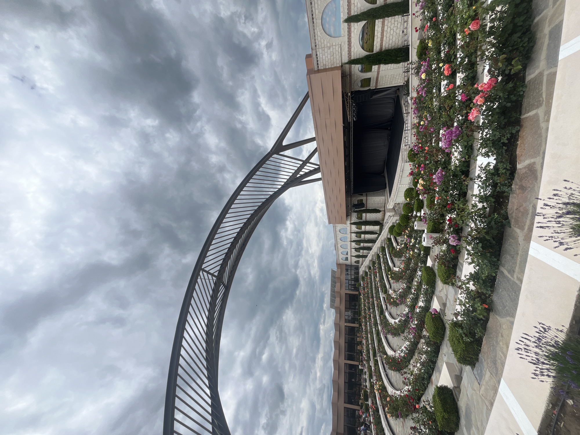 The image depicts an outdoor amphitheater with tiered seating, surrounded by lush rose gardens. The seating areas are curved, with white borders and stone pathways. Rows of blooming roses in various colors, including pink, red, yellow, and purple, are visible in the foreground and on the terraces. In the background, a stage is set against a building with a light-colored stone facade and arched windows, some of which appear to depict painted landscapes. An arched metal structure extends over the seating area towards the stage. The sky is overcast with dramatic, dark clouds. The scene is likely the amphitheater at the Pleven Arch, located near the city of Skobelevo, Bulgaria, a site known for its floral displays and monument. No people are clearly visible interacting with the environment in the image.