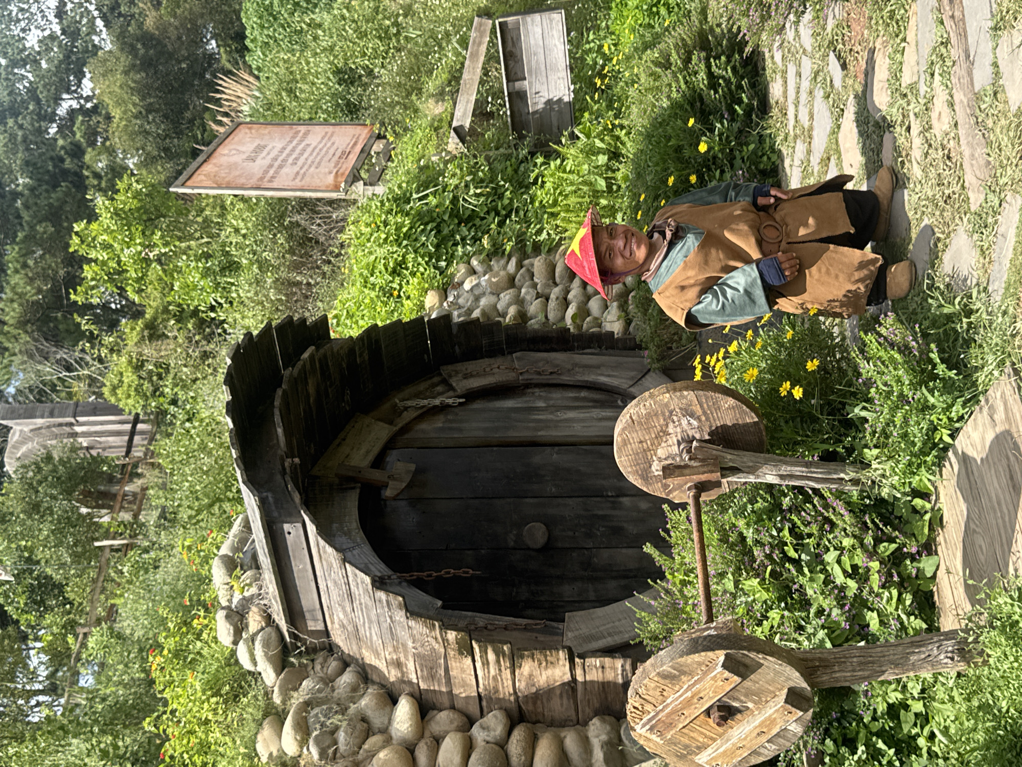 The image depicts a scene at a Hobbit-themed attraction in Thôn 1, Vietnam. In the foreground, a person of short stature, wearing a red conical hat with a yellow star, a green shirt, a brown tunic, and brown shoes, stands on a stone pathway. The person is smiling and facing the camera. Behind the person is a rustic, circular wooden door set into a hillside, constructed from rough-hewn logs and stones. The doorway is partially covered by a wooden roof. To the left of the doorway, there are two large wooden spools on a metal bar, resembling a weightlifting apparatus. The environment is lush and green, with abundant vegetation, including bushes, flowers, and trees. A wooden sign with text is visible in the upper right portion of the image, partially obscured by foliage. The overall impression is of a whimsical, fantasy-inspired setting.