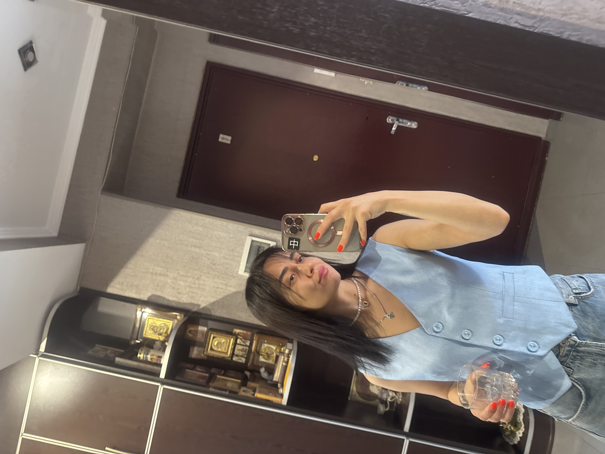 This media file shows a mirror selfie of a young woman with dark hair, wearing a light blue vest and denim shorts. She is holding a clear glass in her right hand and a smartphone in her left. The smartphone screen displays a Chinese character, "中". Her nails are painted bright red. She is wearing a pearl necklace with a small clover pendant.
The background appears to be an indoor setting, likely an apartment hallway or a closet area. In the reflection, a dark brown door with a silver handle and lock is visible. To the left of the door, there's a shelf unit filled with framed religious icons, suggesting a home environment. The overall lighting suggests it is daytime. The woman's expression is playful, with her lips pursed. The setting and the presence of religious icons may hint at the location being in or around a religious community, or simply a personal collection. The mirror itself has a dark frame and reflects the ceiling, which has a light fixture. The scene conveys a casual, everyday moment.