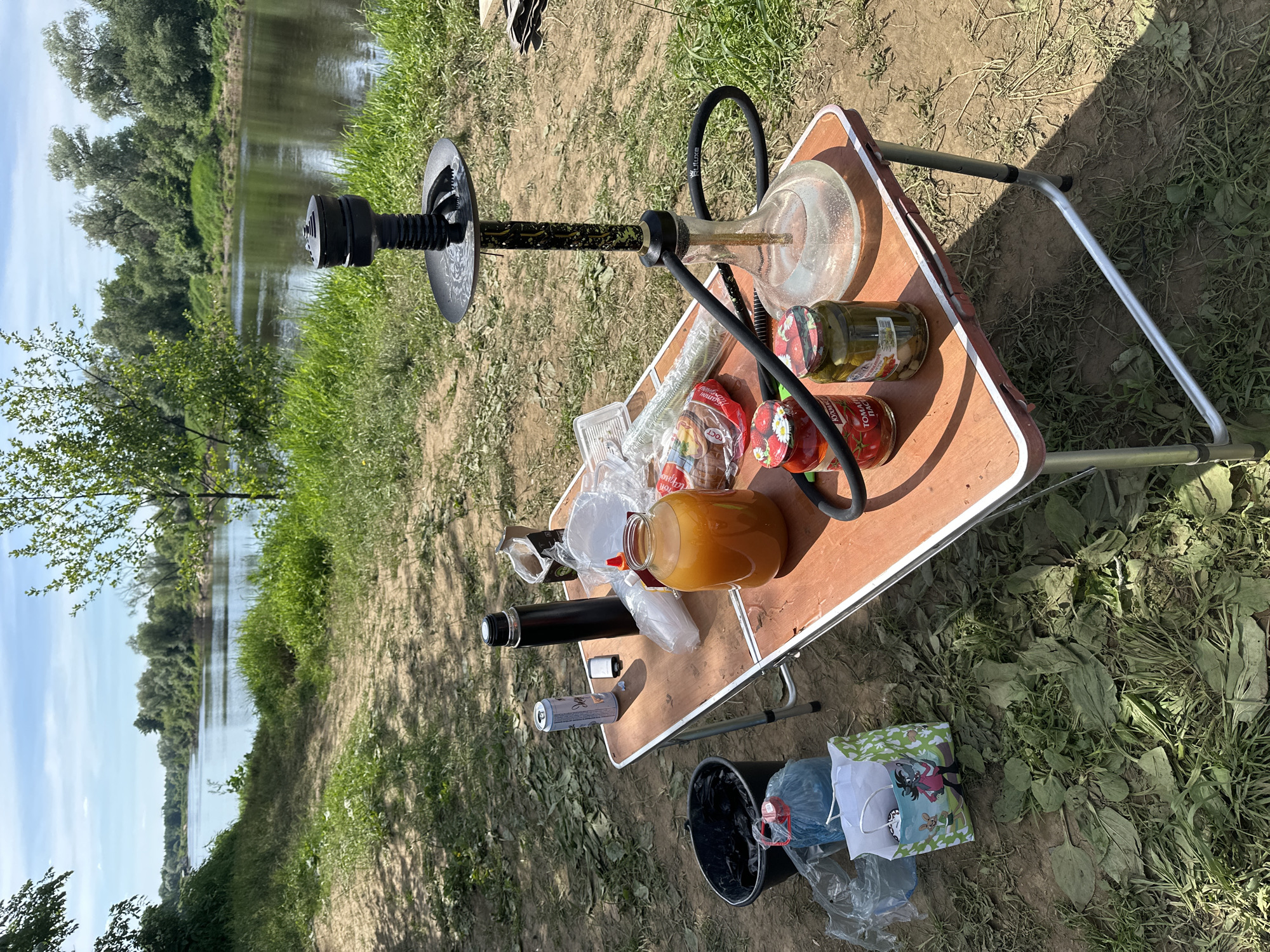 The image depicts an outdoor leisure setup positioned on a dirt path next to a river, consistent with a natural area in or near Ladoga, Russia. In the foreground, a portable rectangular folding table with a reddish-brown top and silver metal legs is placed on dry, earthy ground with sparse green grass and scattered dry leaves.
On the table, from left to right, are various items: a silver aluminum can, a black thermos, a small white object (possibly a lighter or small battery pack), several clear plastic bags containing food items such as bread, a large clear plastic bottle filled with an orange liquid, and two glass jars with metal lids containing red tomato paste and green pickled cucumbers, respectively. A black hookah with a clear glass base filled with water, a black hose, and a black bowl/tray is prominently displayed on the right side of the table.
To the left of the table, on the ground, a black plastic bucket contains a black trash bag. Adjacent to the bucket are a clear plastic bag holding a blue object and a small light green paper gift bag featuring cartoon characters.
The midground features a wide, calm river flowing horizontally across the frame, reflecting the bright blue sky with scattered white clouds. The riverbanks are heavily vegetated with dense green trees and bushes, indicating a lush natural environment. A dirt path leads down towards the river on the near bank. The scene is illuminated by direct sunlight, casting distinct shadows. No people are visible in the image.