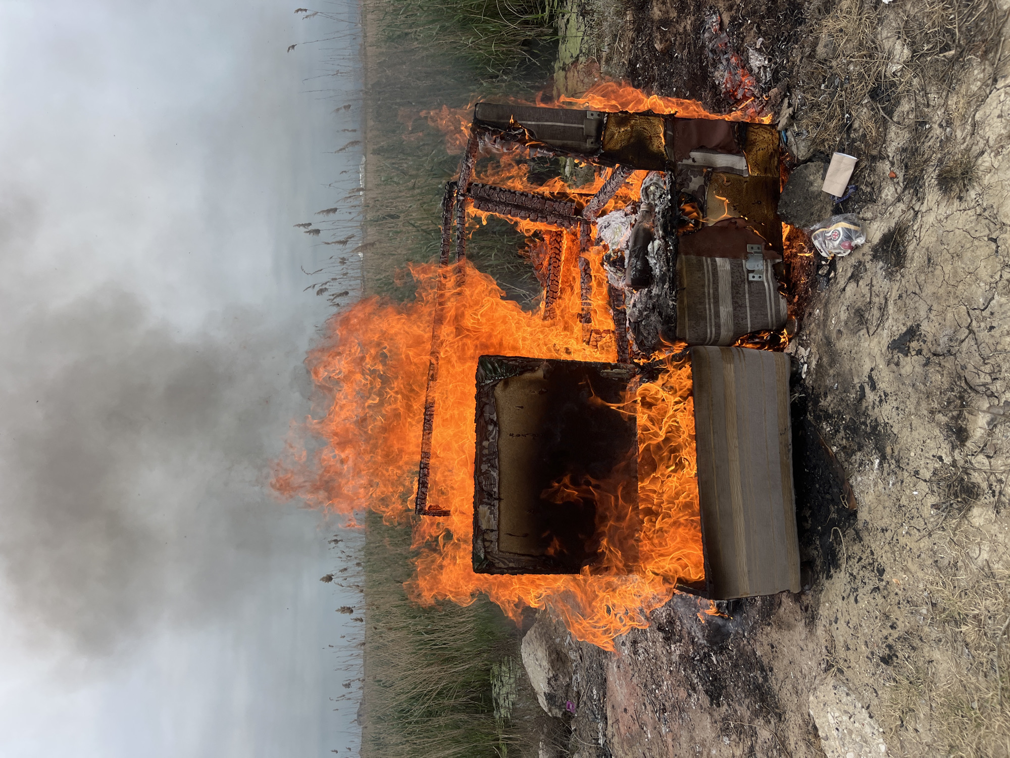 A fire is burning an object, potentially furniture, in a field near Kaspiysk, Russia. Flames engulf what appears to be a frame structure and cushioned objects. Gray smoke rises into a gray sky. Dry reeds are visible in the background, and barren ground surrounds the fire. Some trash and debris are scattered nearby.