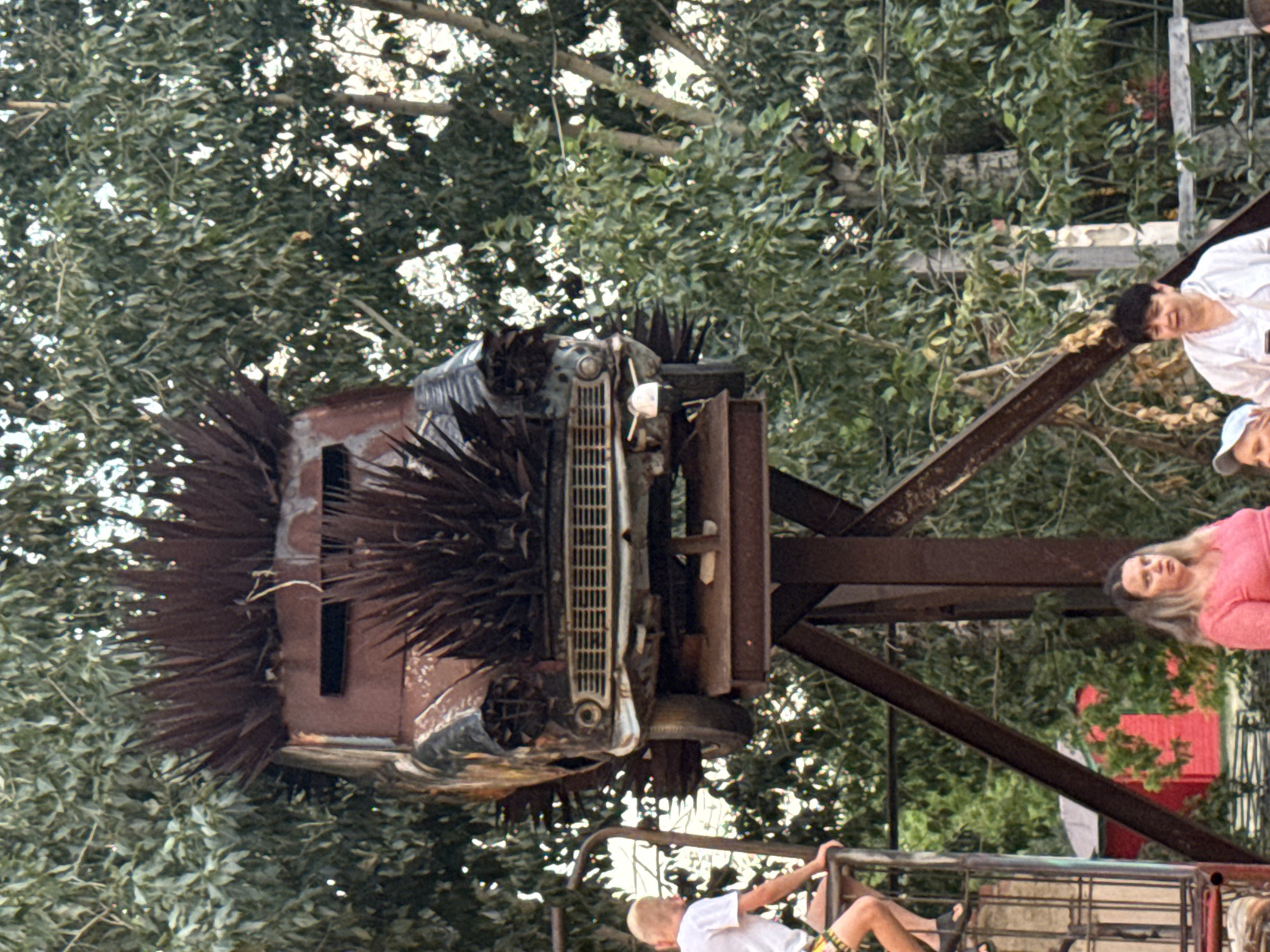An unusual art installation is depicted, featuring a vintage car mounted on a metal structure, adorned with spiky metal protrusions. The car appears weathered and rusted, with its front end prominently displayed. The installation is set against a backdrop of lush green trees. In the foreground, several individuals are visible. A young boy with blond hair is positioned on a metal platform to the left, looking towards the installation. To the right, two women are standing, one wearing a pink top and the other a white top. A red building is partially visible in the background behind the metal structure. The scene appears to be an outdoor public space, likely part of an art park or festival in the city of Yarovoye, Russia.