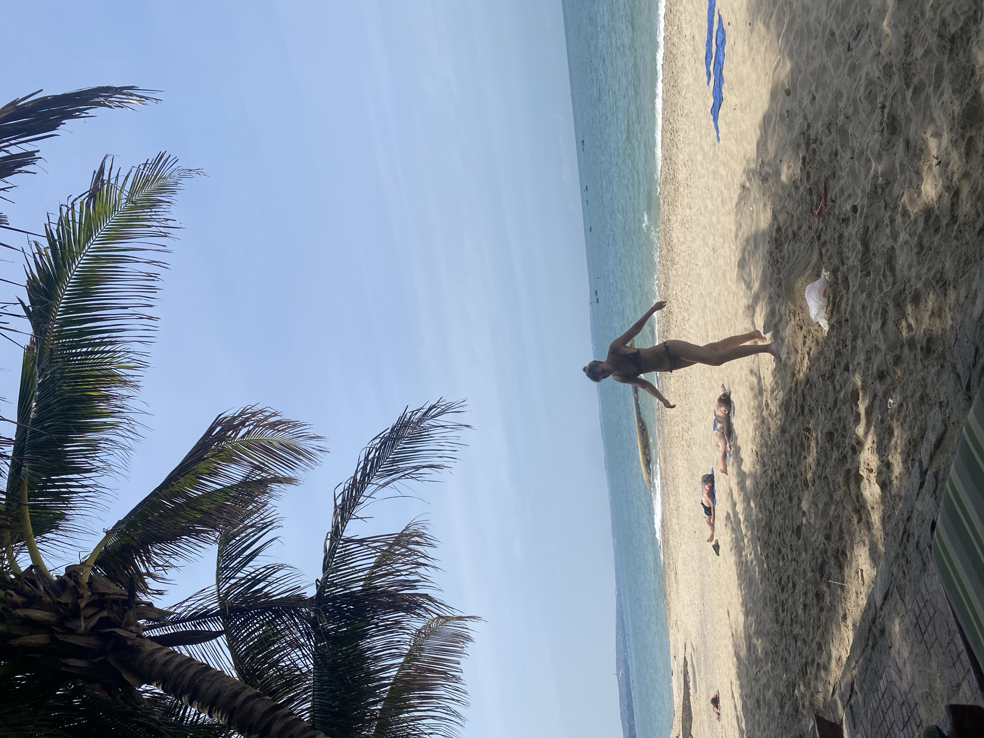 A full shot captures a sunny day at the beach in Phu Thuy Ward, Vietnam. The scene is dominated by the sandy shore, with the turquoise ocean meeting the horizon under a clear blue sky. In the foreground, a lush palm tree casts shadows on the sand, its fronds reaching towards the sky. A woman in a bikini walks across the beach, her body in mid-stride, facing away from the camera. Several other individuals are scattered across the sand, some lying down, appearing to sunbathe. Blue towels are placed on the sand near the water's edge. In the distance, several boats are visible on the water. Trash, including a white plastic bag and some red debris, is present on the sand.
