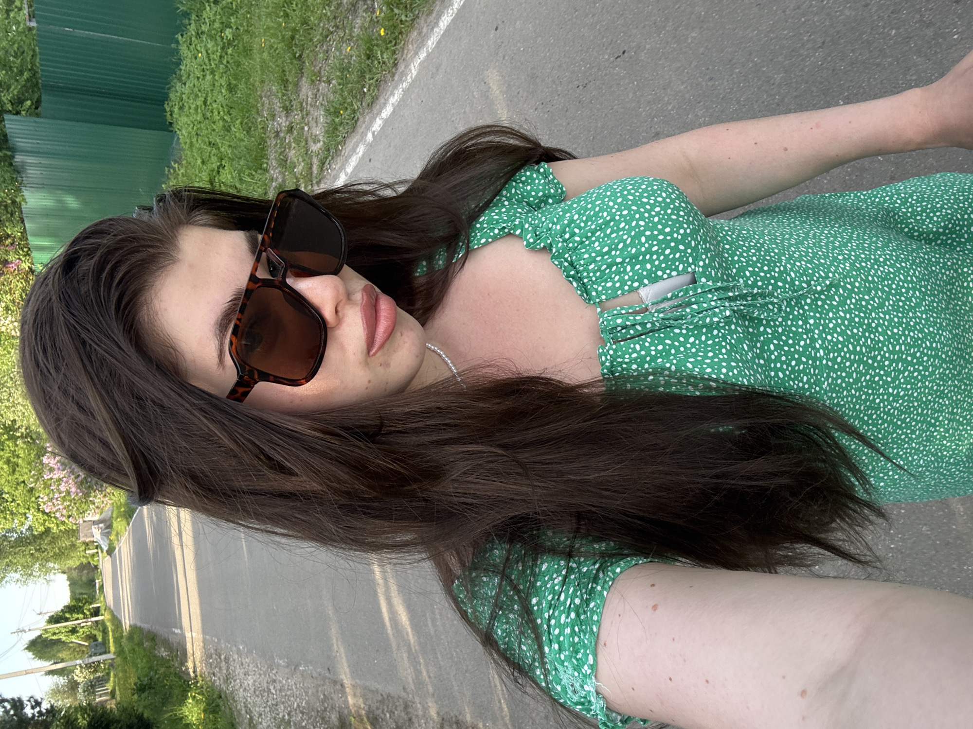 A young woman with long, dark brown hair, wearing sunglasses and a green and white polka-dot dress, is standing on a paved road. The road extends into the distance, flanked by green vegetation and a green fence on the left. The photograph appears to be taken in a rural area, possibly in the vicinity of Мещёры, Russia. The image seems to be a selfie taken outdoors during daylight hours.