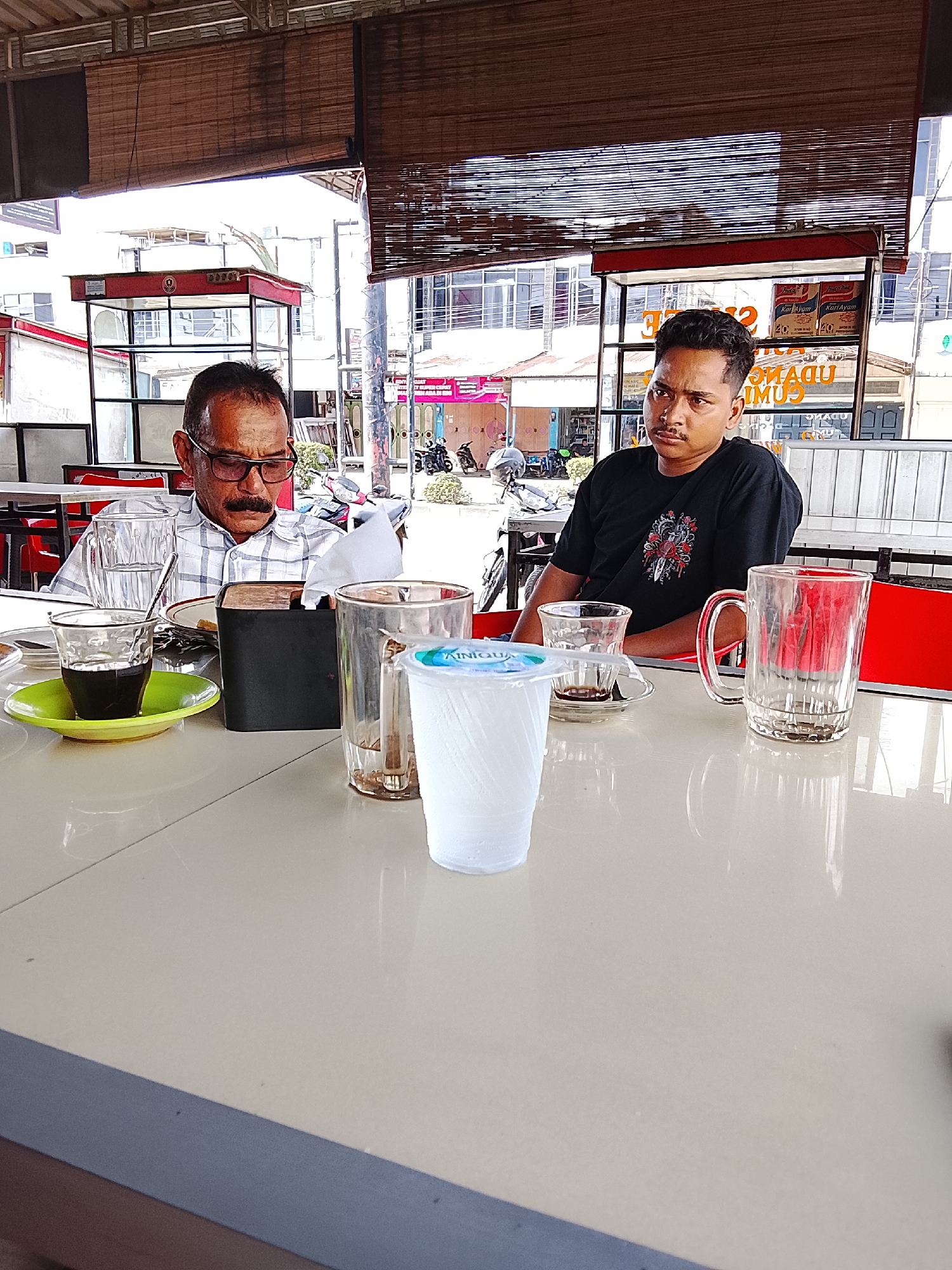 Two men are seated at a light-colored table in an outdoor cafe setting. The older man on the left wears glasses and a mustache. He is dressed in a white and blue patterned shirt. The younger man on the right wears a black t-shirt. Several glasses and a cup of coffee are on the table. There is also a napkin holder. The location appears to be Lhokseumawe, Indonesia, based on visual cues like signage, the style of the buildings, and the presence of scooters on the street in the background. Beyond the cafe, there is a street with vehicles and buildings. A partially retracted awning is visible above the patrons.