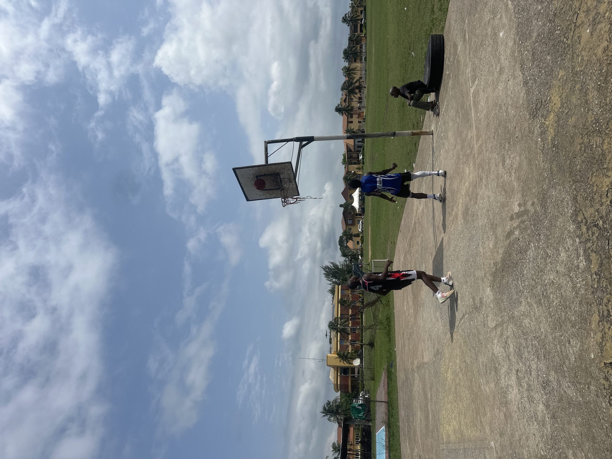 In Ajara, Nigeria, on a concrete basketball court, three individuals are present. Two young men are actively engaged in a game of basketball. One, wearing a black and red jersey and white shorts, stands near the foreground, facing away from the viewer. The other, dressed in a blue jersey and black shorts, is positioned closer to the basketball hoop, with his back to the camera. A third individual, a young man in a black t-shirt and dark pants, sits on a large black tire to the right of the basketball hoop, observing the game. The basketball hoop itself is weathered, with a net that appears to be torn or absent. The court is surrounded by a chain-link fence. Beyond the court, a grassy field extends, with several buildings and palm trees visible in the background under a cloudy blue sky. A swimming pool is situated to the left of the basketball court.