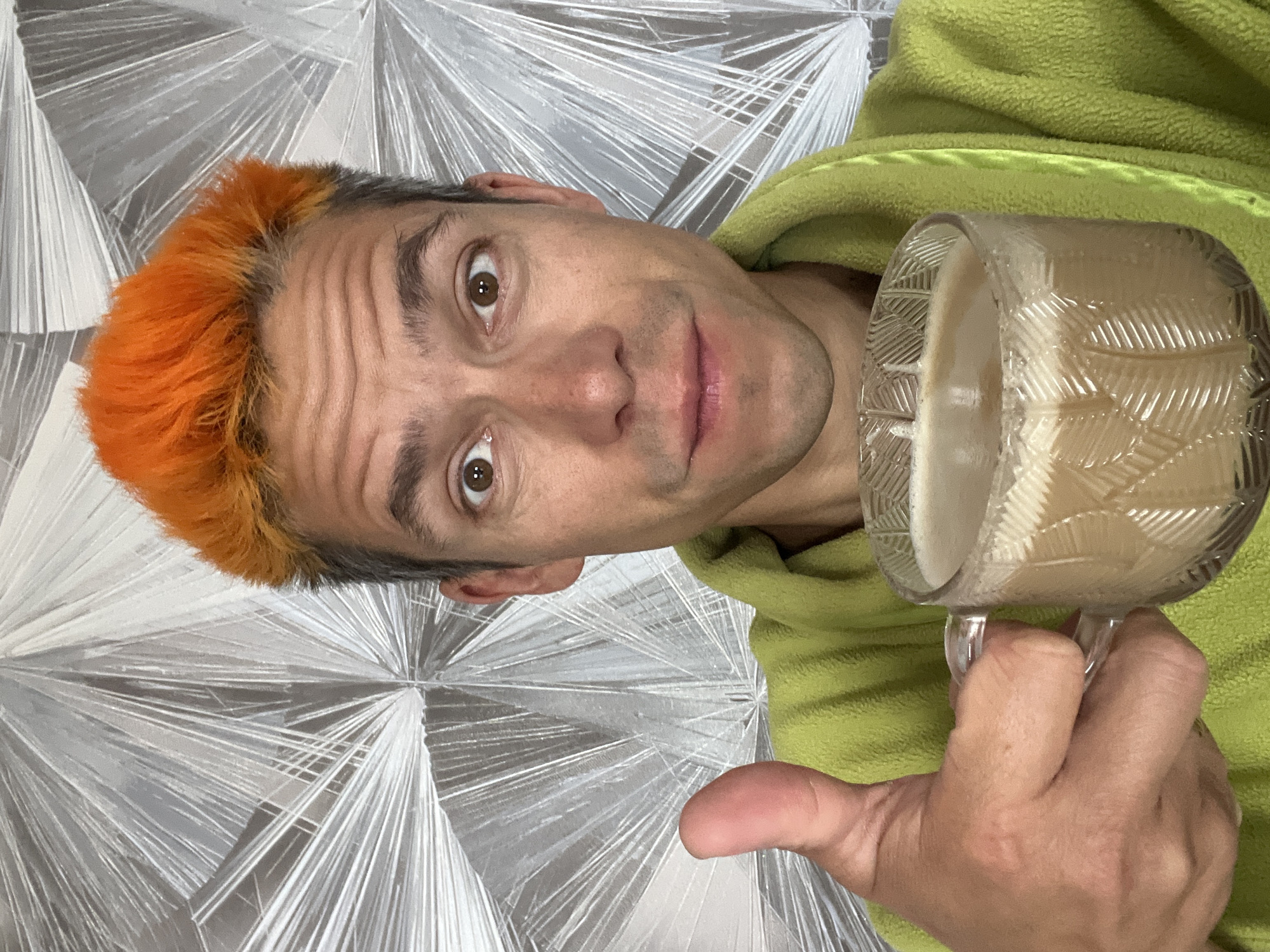 A man with brightly dyed orange hair and a light green robe is holding a mug of coffee and giving a thumbs-up. He is standing in front of a patterned silver wallpaper. The image does not provide enough information to determine a location in the city of Chaikovsky, Russia.