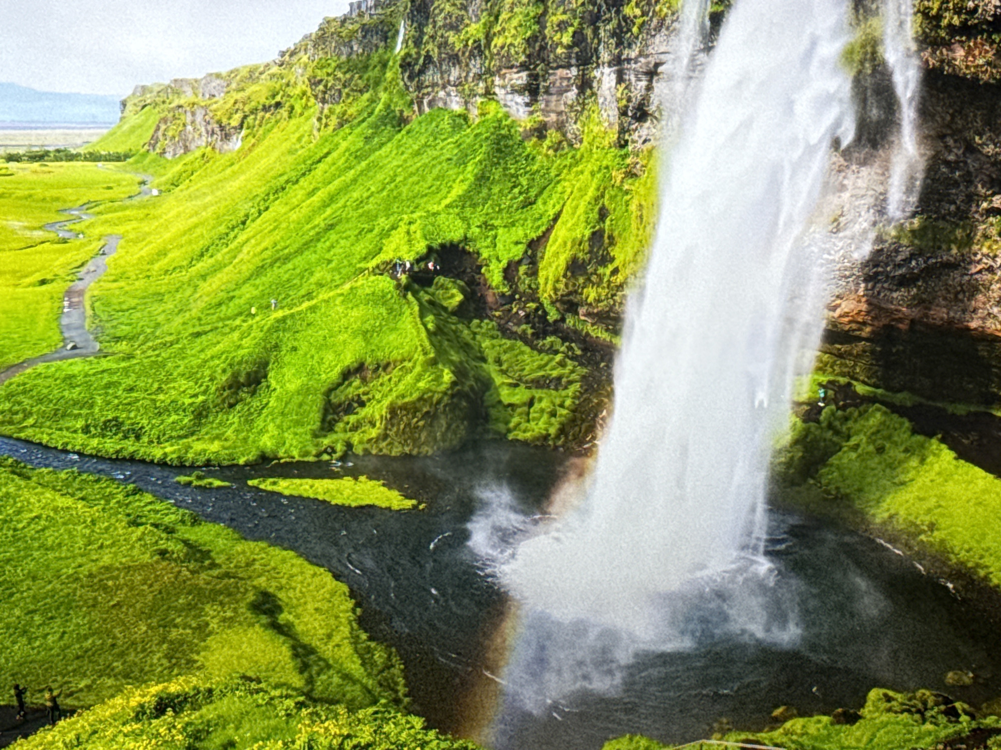 A scenic view captures a majestic waterfall cascading down a lush green mountainside. The water plunges into a dark pool below, creating a misty spray that catches the sunlight, forming a faint rainbow. A winding path, possibly a trail for tourists, snakes its way up the grassy slope. Several small figures, likely hikers, can be seen on the path, adding a sense of scale to the grand landscape. The surrounding environment is characterized by vibrant green vegetation covering the steep terrain, with rocky outcrops and the sheer cliff face of the waterfall providing a dramatic backdrop. The sky above is overcast, casting a soft, diffused light over the scene. The image is taken from an elevated perspective, offering a wide view of the natural beauty. This natural landmark is not located in Burgas, Bulgaria.