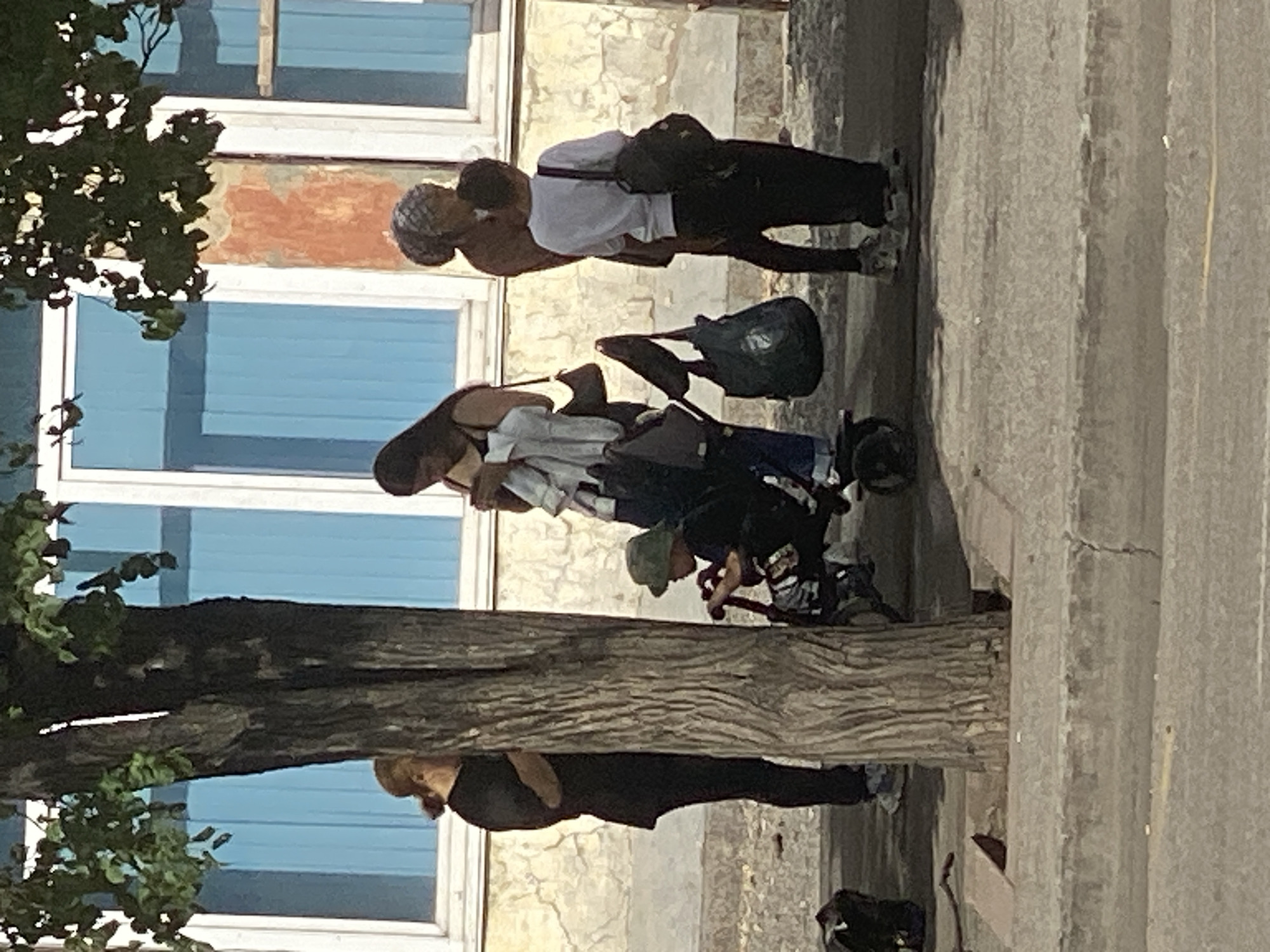 A group of people are gathered near a tree on a sidewalk in front of a building. A young child wearing a bucket hat is sitting in a stroller. To the left of the child, a woman in a black shirt and pants stands behind the tree. To the right of the child, two adults are standing, one holding a bag and the other wearing a white shirt and backpack. The building behind them has blue windows and weathered walls. The scene appears to be outdoors in an urban environment.