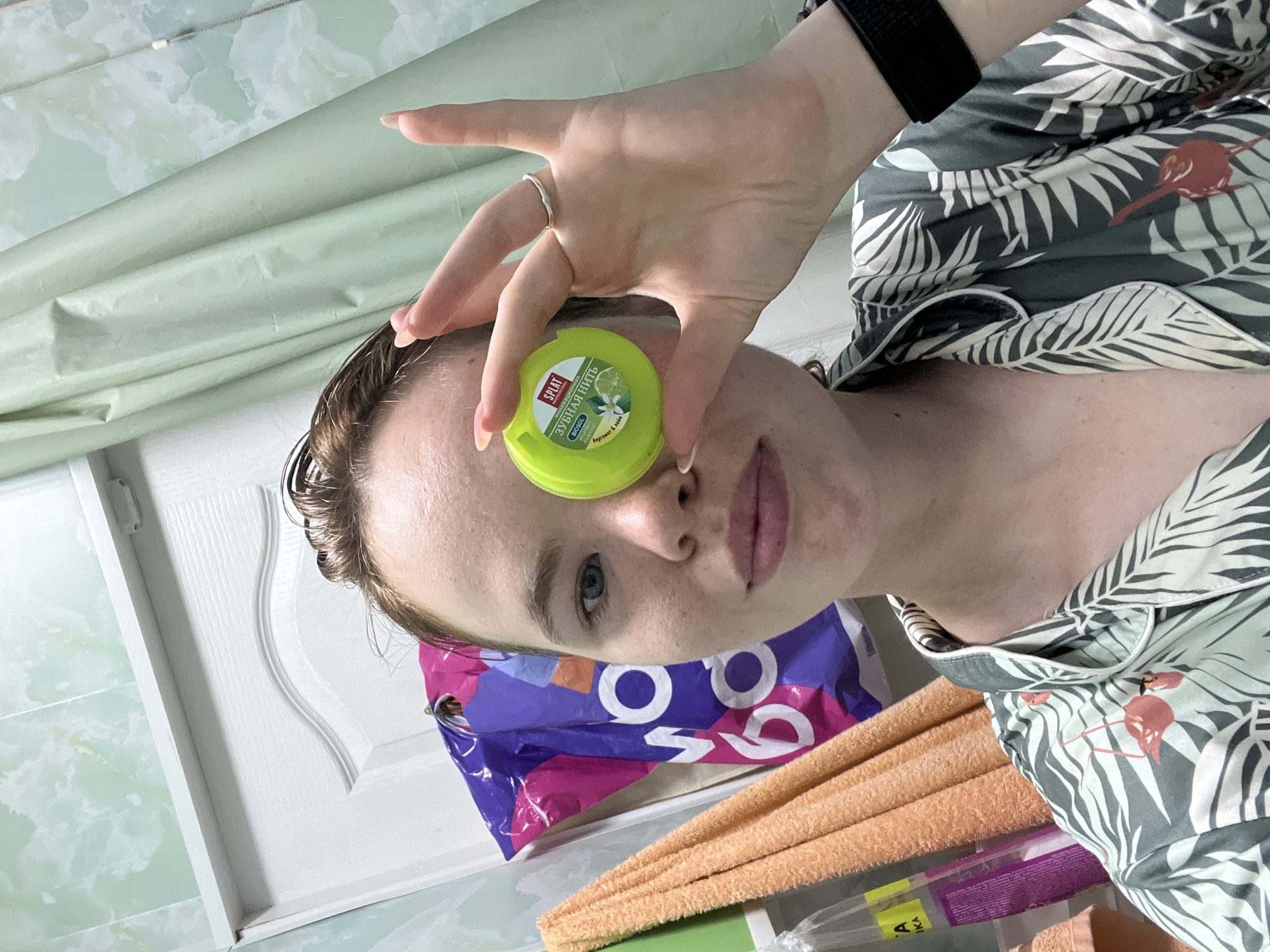 A young Caucasian woman with short, wet, brown hair and light blue eyes is shown from the chest up, holding a small, green, round container of "Splat" brand dental floss in front of her right eye. The container has text in Russian and a depiction of a lime and a flower. She is wearing a gray and green patterned pajama top with a tropical leaf and flamingo design. A silver ring is on her left ring finger. In the background, a light green, patterned shower curtain is visible, along with a white door and a rack with orange and purple towels or bags. A black wristband is on her left wrist. The lighting suggests an indoor bathroom setting. The location in Chaikovsky, Russia is not discernible from the image itself.