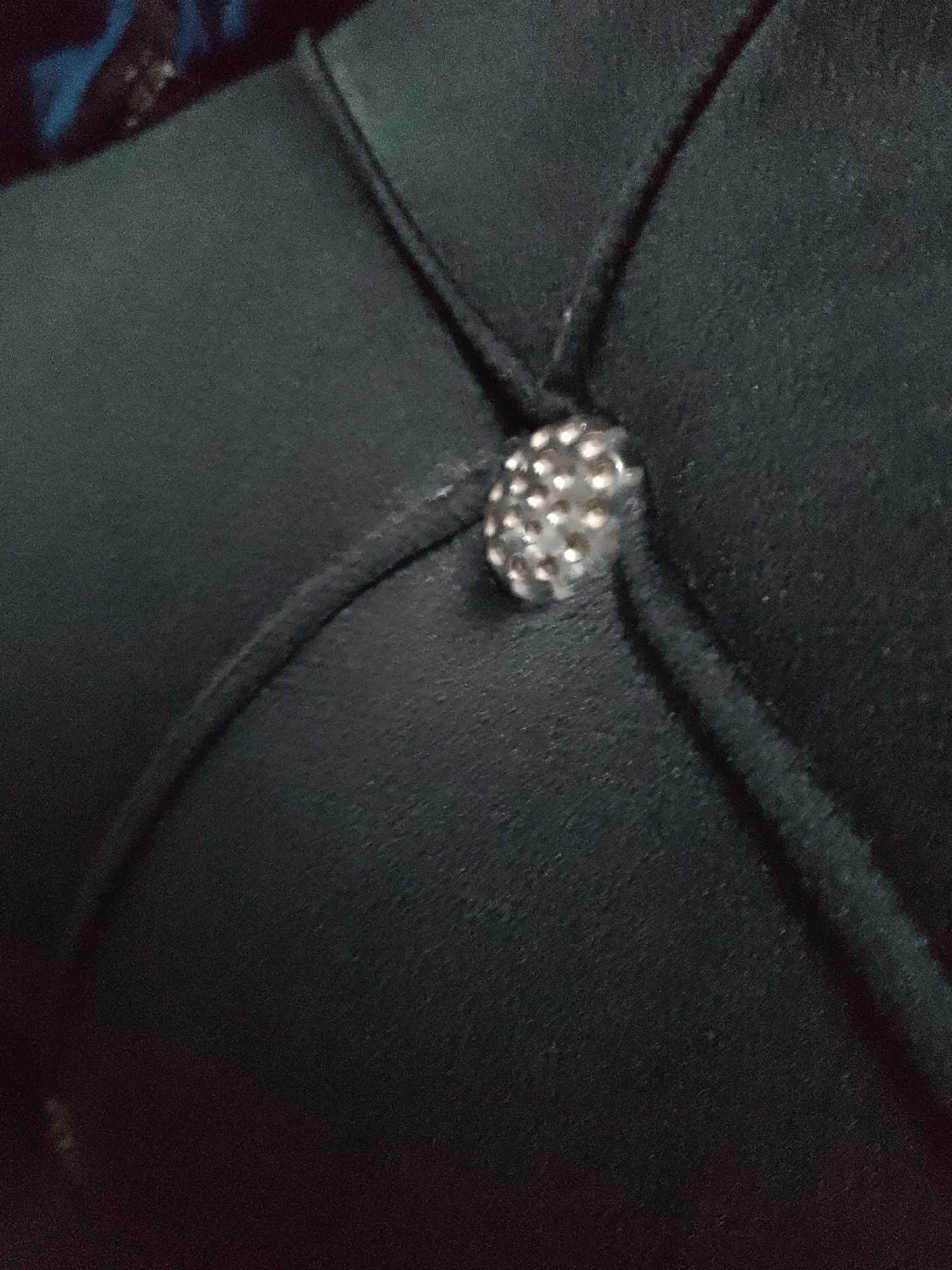 The image is a close-up, slightly out-of-focus shot of a dark green upholstered surface, possibly furniture. A single, small, spherical embellishment, appearing to be a rhinestone or sequin-covered button, is centrally located. The upholstery has tufting, indicated by the indentations and stitching lines that form a diamond pattern around the embellishment. The lighting is dim, with some highlights on the button and stitching. The background is largely out of focus and dark, with a hint of a blue and dark pattern visible in the upper left corner, suggesting another object or surface. There are no people visible in the image. The image does not contain any elements that would indicate its location in Jauro Yinu, Nigeria.