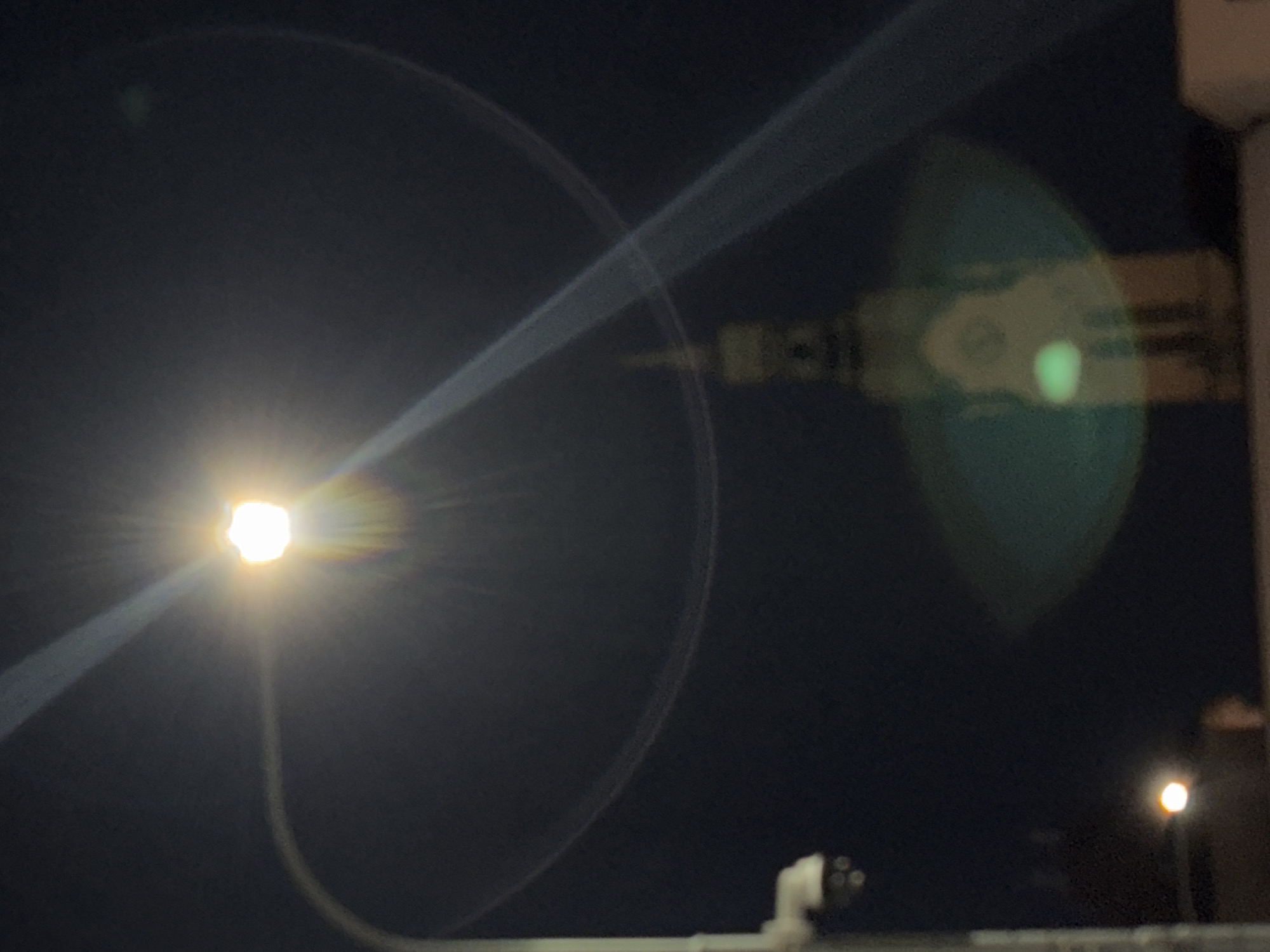 An out-of-focus, night time, outdoor photograph shows a street light on a pole with a security camera attached to the side of the pole. The street light emits a bright glow with lens flare, creating arcs and streaks of light in the dark sky. In the background, a blurry, tall, light-colored building resembling a church tower with a clock face is visible. There are other out-of-focus lights in the lower part of the image. The overall impression is a nighttime urban scene.