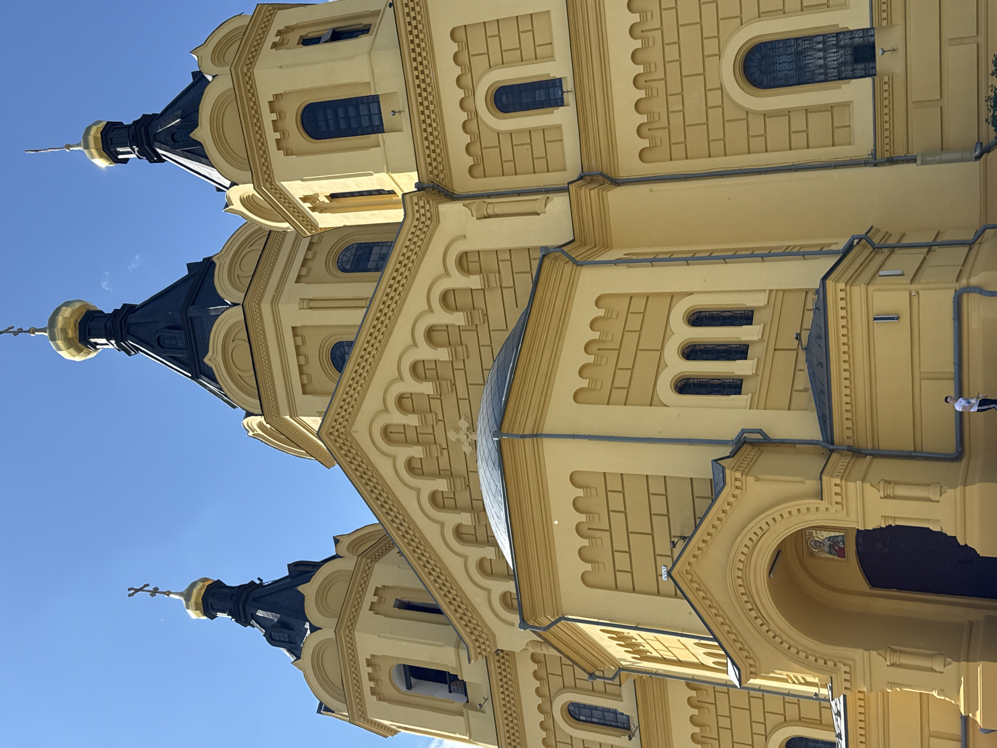 This is a low-angle, full-shot photograph of the exterior of the Alexander Nevsky Cathedral, located in Nizhny Novgorod, Russia. The cathedral is predominantly yellow with dark blue roofs and gold domes topped with crosses. Multiple turrets and domes are visible, as are arched windows with dark grates and decorative brickwork. A small icon is visible above the main arched entrance. A single person in a white shirt and dark pants is walking away from the cathedral on the right side of the image. The sky is clear and blue.