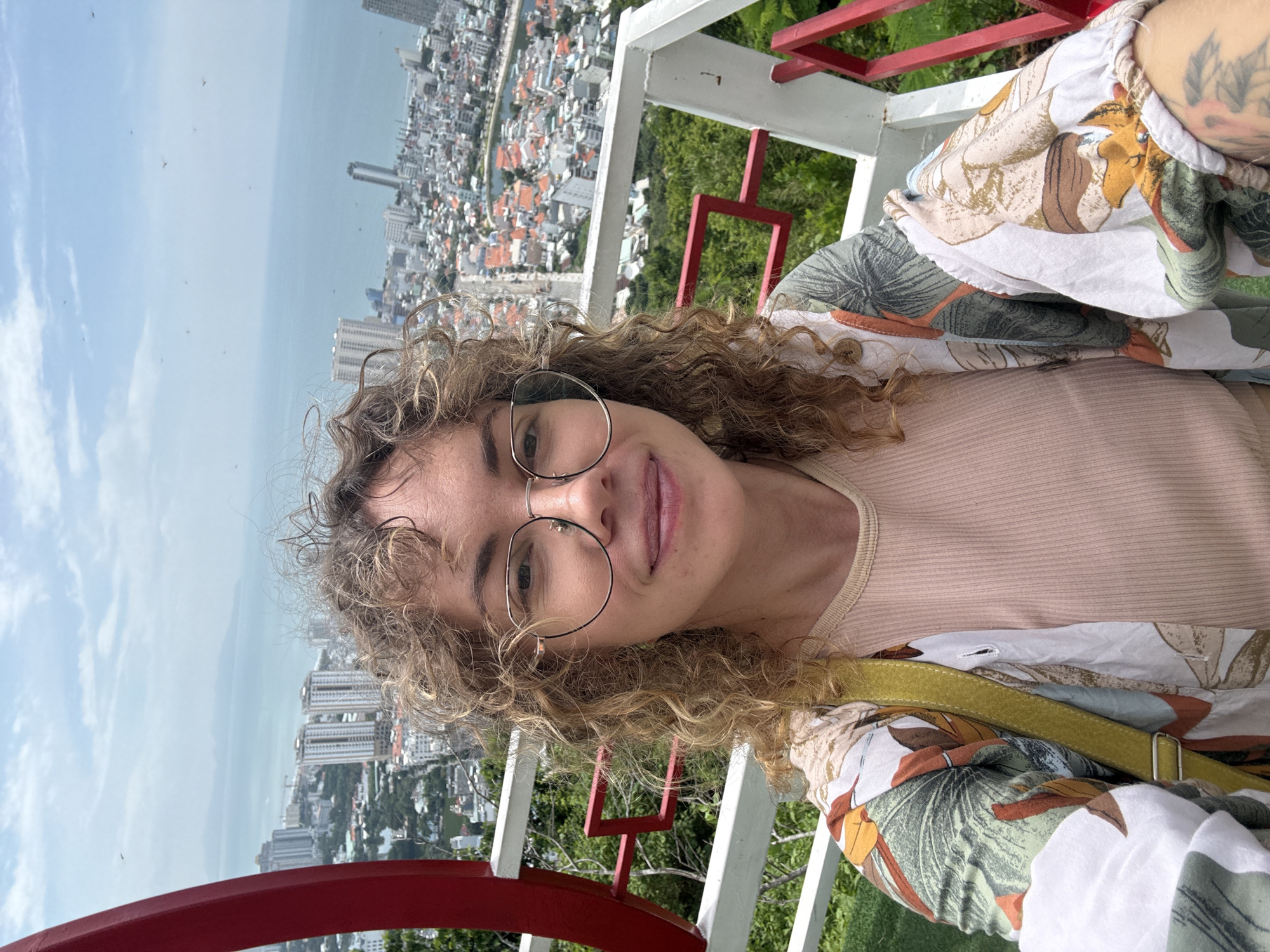 A woman with curly brown hair and glasses is taking a selfie on a balcony overlooking a cityscape. She is wearing a light pink ribbed top and a colorful floral print kimono-style jacket. Behind her, a dense city with many buildings and a body of water can be seen. The sky is partly cloudy. This appears to be the Linh Phong Pagoda in Ho Chi Minh City, Vietnam, known for its panoramic views of the city and the sea.