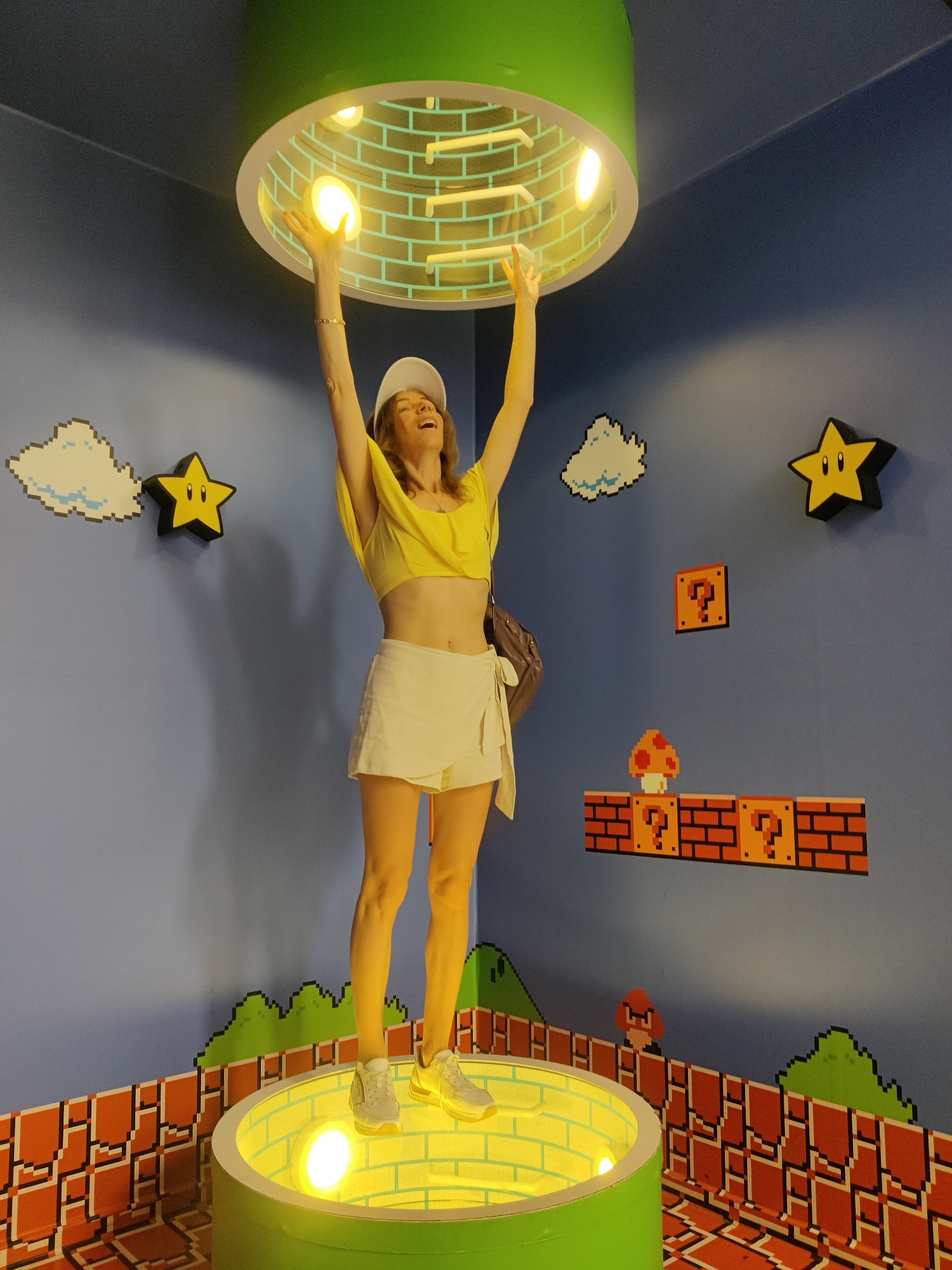 In Thessaloniki, Greece, a woman stands on a cylindrical platform, her arms raised toward a circular, illuminated ceiling fixture. The scene is decorated with pixelated graphics reminiscent of the Super Mario video game franchise. The walls feature images of clouds, stars, question mark blocks, and a mushroom. The woman is wearing a yellow crop top, a white skirt, a white cap, and white sneakers. She has a brown bag slung over her shoulder. The platform she is standing on is green and lit from within by a yellow light. The floor is also decorated with pixelated brick patterns.