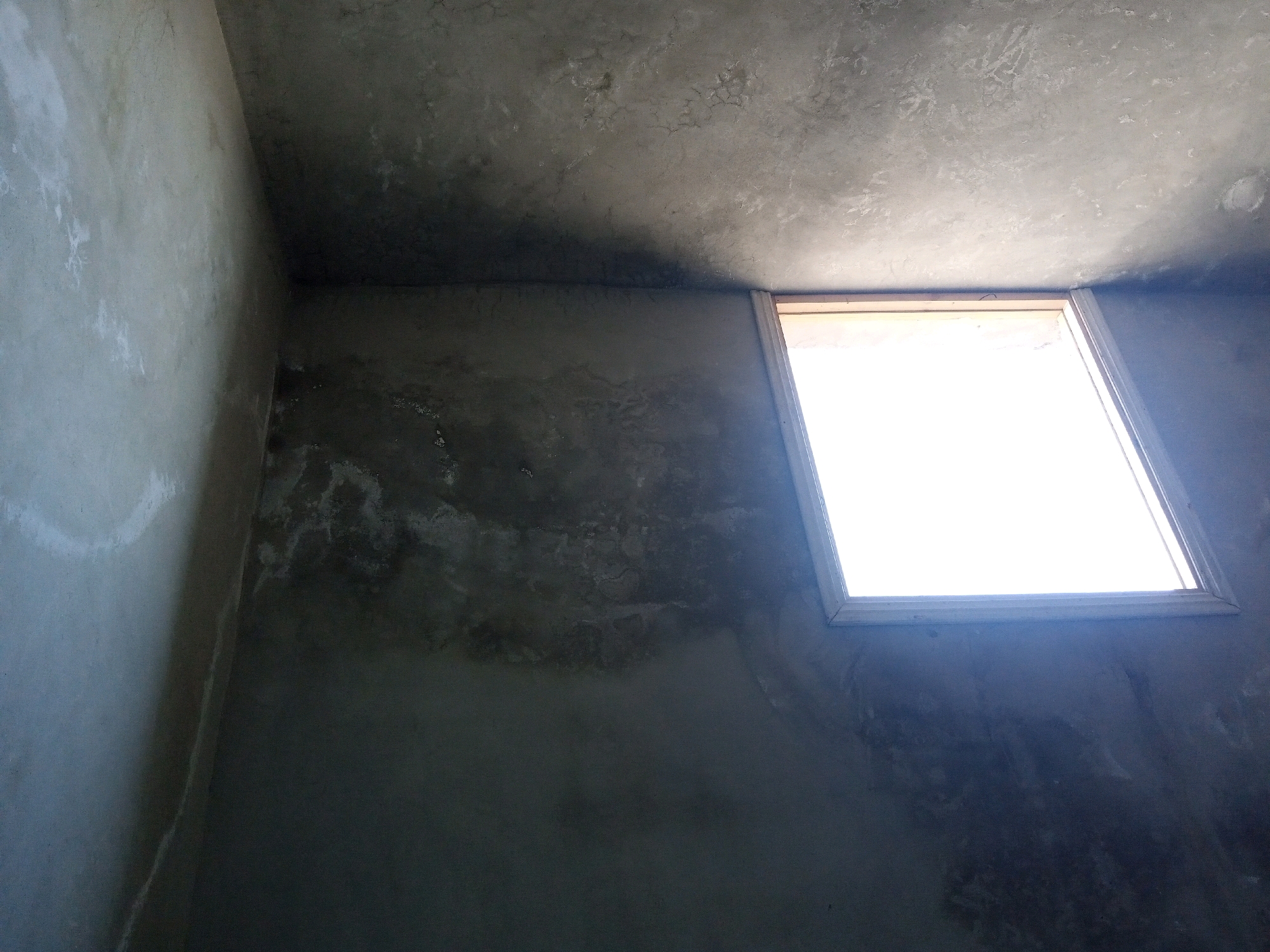 The image shows the interior of a dimly lit room with unfinished concrete walls and ceiling. A rectangular window, letting in a bright light, is visible in the upper right quadrant of the image. The walls exhibit uneven textures and some discoloration, suggesting a construction or renovation phase. The overall environment appears to be an indoor space within a building, potentially under construction or a recently completed structure. There are no people or discernible objects present in the scene other than the structural elements and the window. The lighting conditions are characterized by a strong source of light from the window contrasting with the shadowed areas of the room.