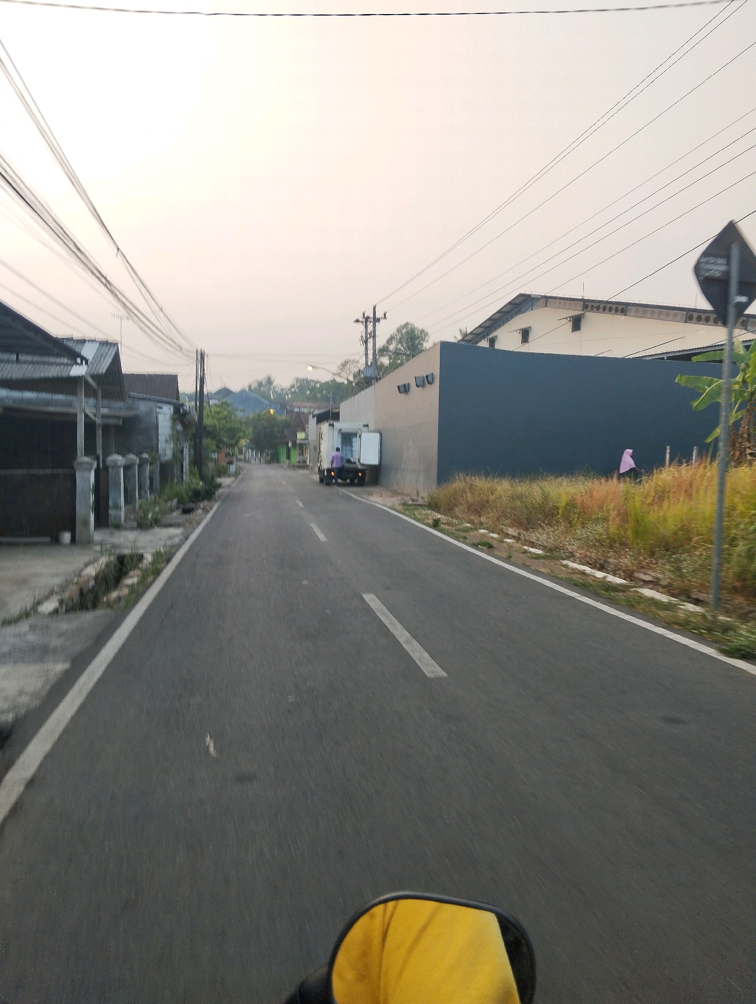 The image depicts a view from a moving vehicle on a paved road in Salatiga, Indonesia. The road is lined with houses on the left side and a large building with a dark gray wall on the right. The sky is overcast and appears to be early morning or late afternoon.
On the left side of the road, there are several houses with corrugated metal roofs. Some houses have fences and pillars in front of them. Vegetation is present along the side of the road and in front of the houses.
On the right side of the road, a long, dark gray wall dominates the foreground, with dry grass and some greenery growing in front of it. A person wearing a pink headscarf is visible walking near the wall. Further down the road, a white truck with its rear doors open is parked, and a person in a purple shirt is standing next to it.
Power lines stretch across the sky. The road has a white line marking the center and a white line marking the edge on the left. The image is captured from the perspective of someone on a motorcycle, as evidenced by the reflection of a yellow object (likely a jacket or seat) in the rearview mirror at the bottom of the frame. The overall scene suggests a residential or mixed-use area in a town or city.