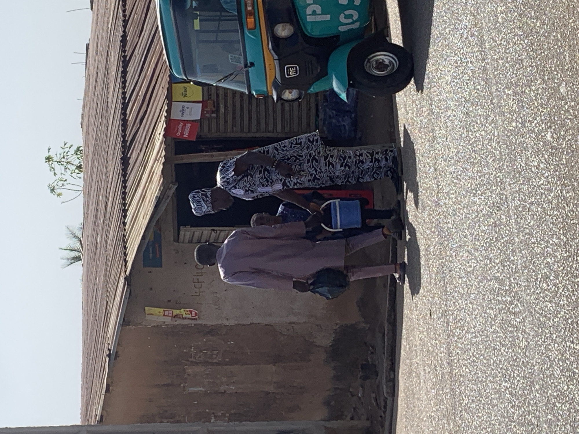 The image depicts a street scene in what is identified to be Jos, Nigeria. A turquoise-colored motorized tricycle is parked near the side of a building. A woman wearing a black and white patterned garment and head covering is standing next to the tricycle. A man in a lavender-colored outfit walks towards the tricycle, carrying a dark bag or purse. A child is positioned between the man and woman. The building alongside the group appears to be a store or shop, with various product posters visible. The building has a corrugated metal roof, and the paint on the walls is faded and worn. The street is paved.